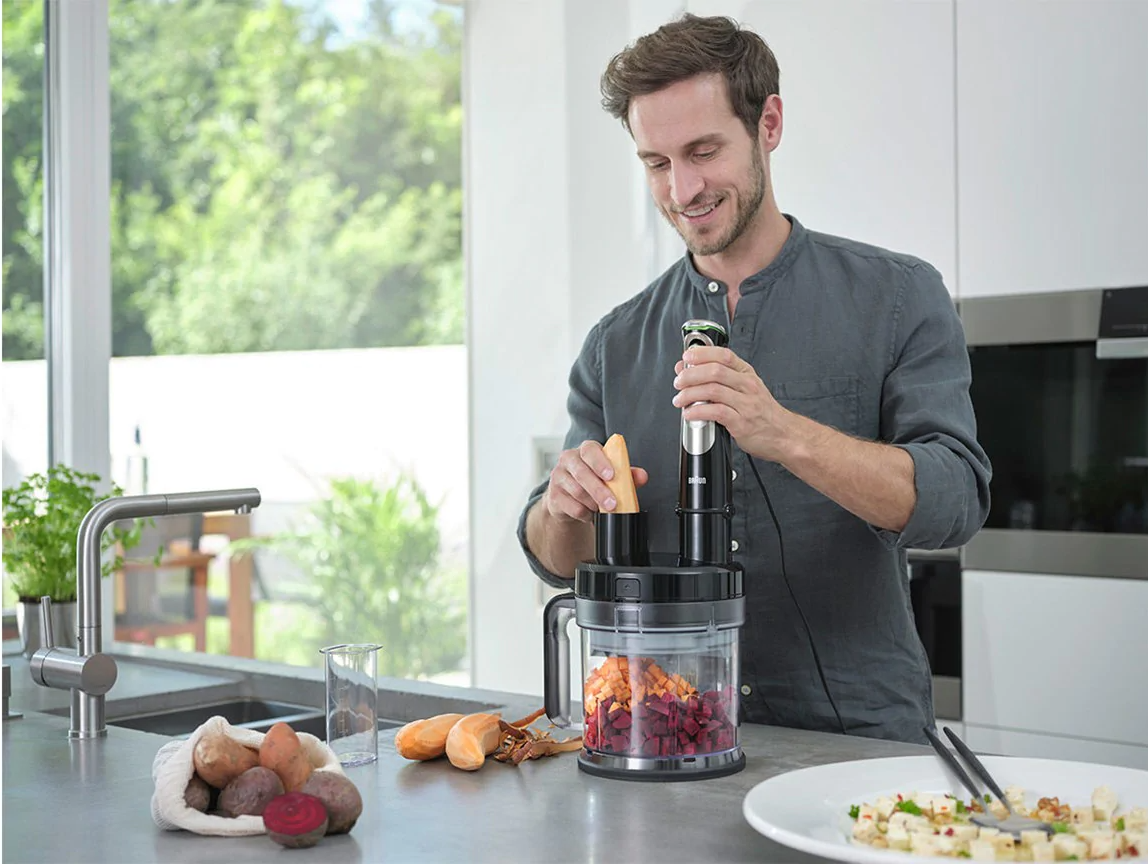 Un homme prépare de la nourriture avec un mixeur plongeant. Il y a des légumes dans un récipient.