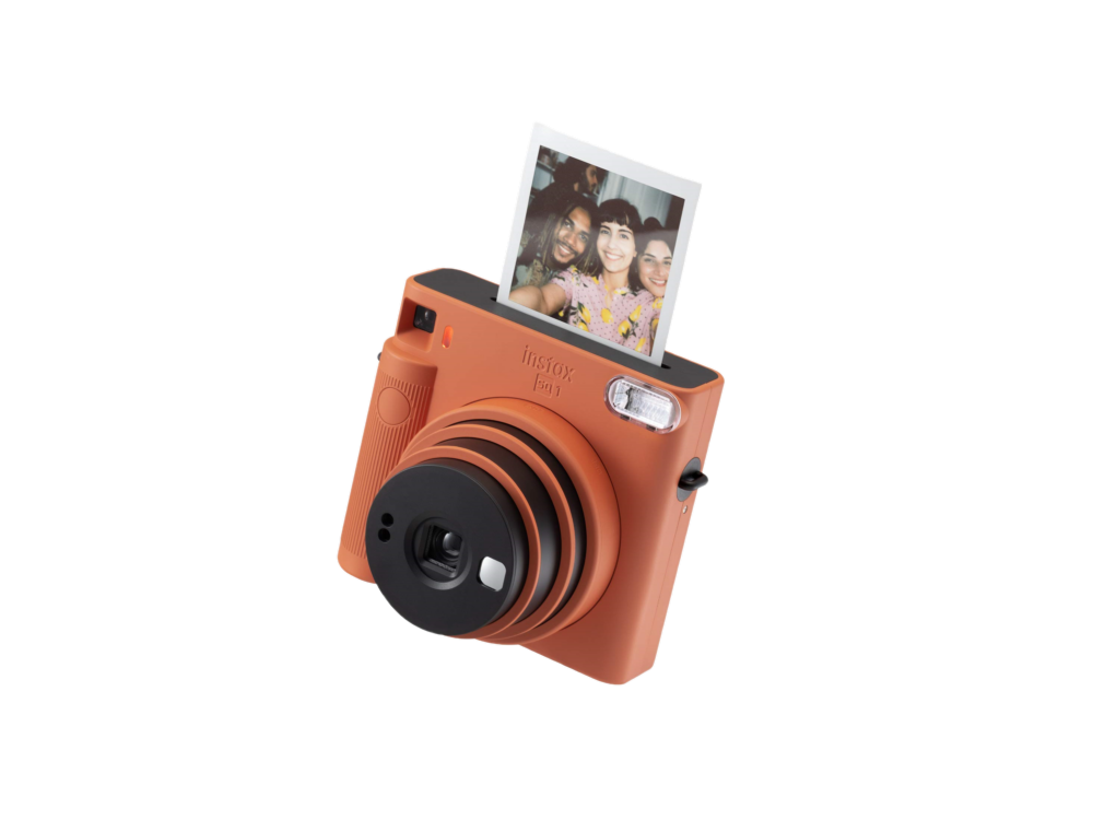 Una cámara Instax naranja con una foto que se expulsa, lente negra y fondo negro.