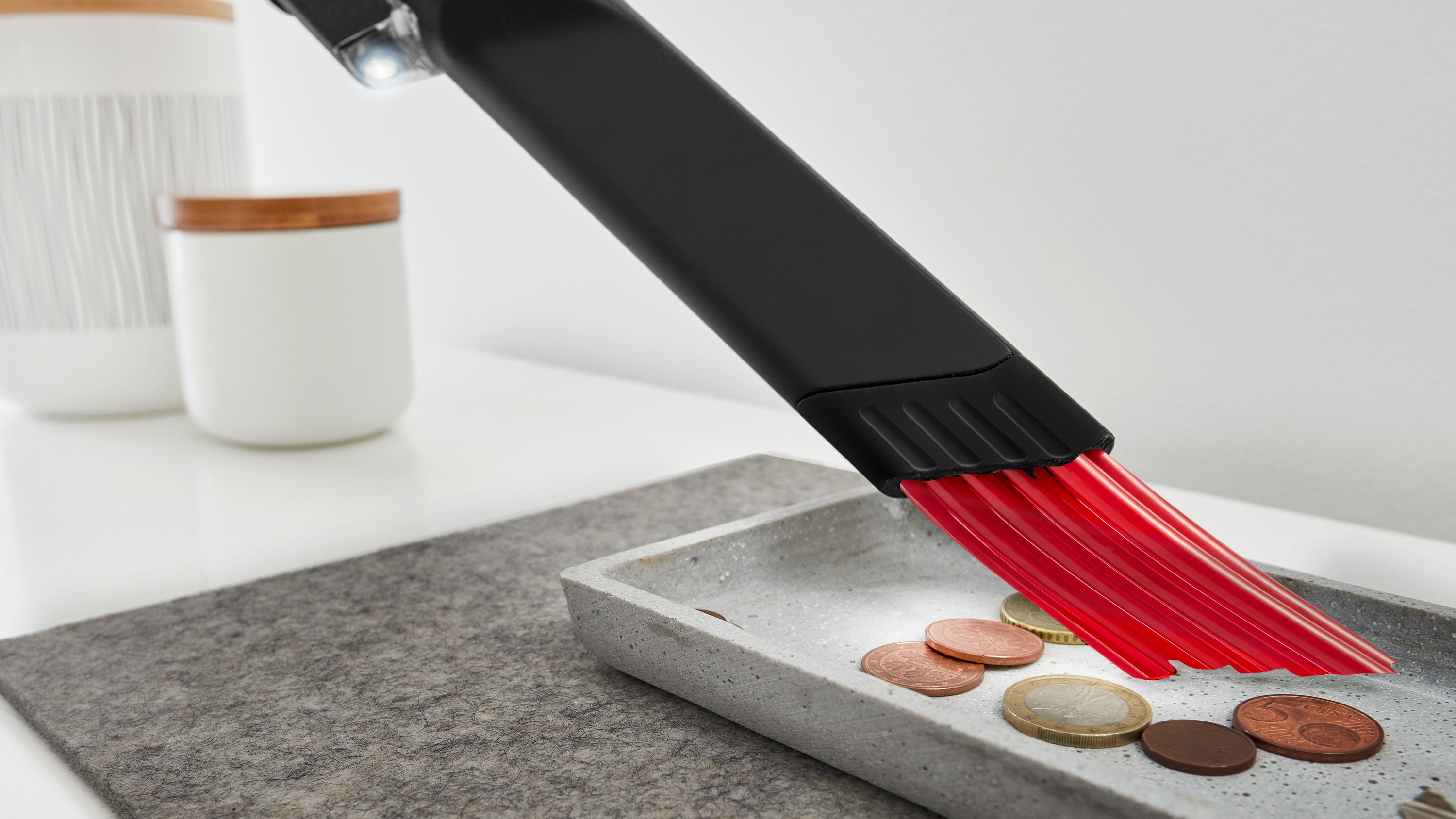 Staubsauger-Bürstenkopf reinigt ein Tablett mit Münzen. Rote Borsten sind sichtbar.