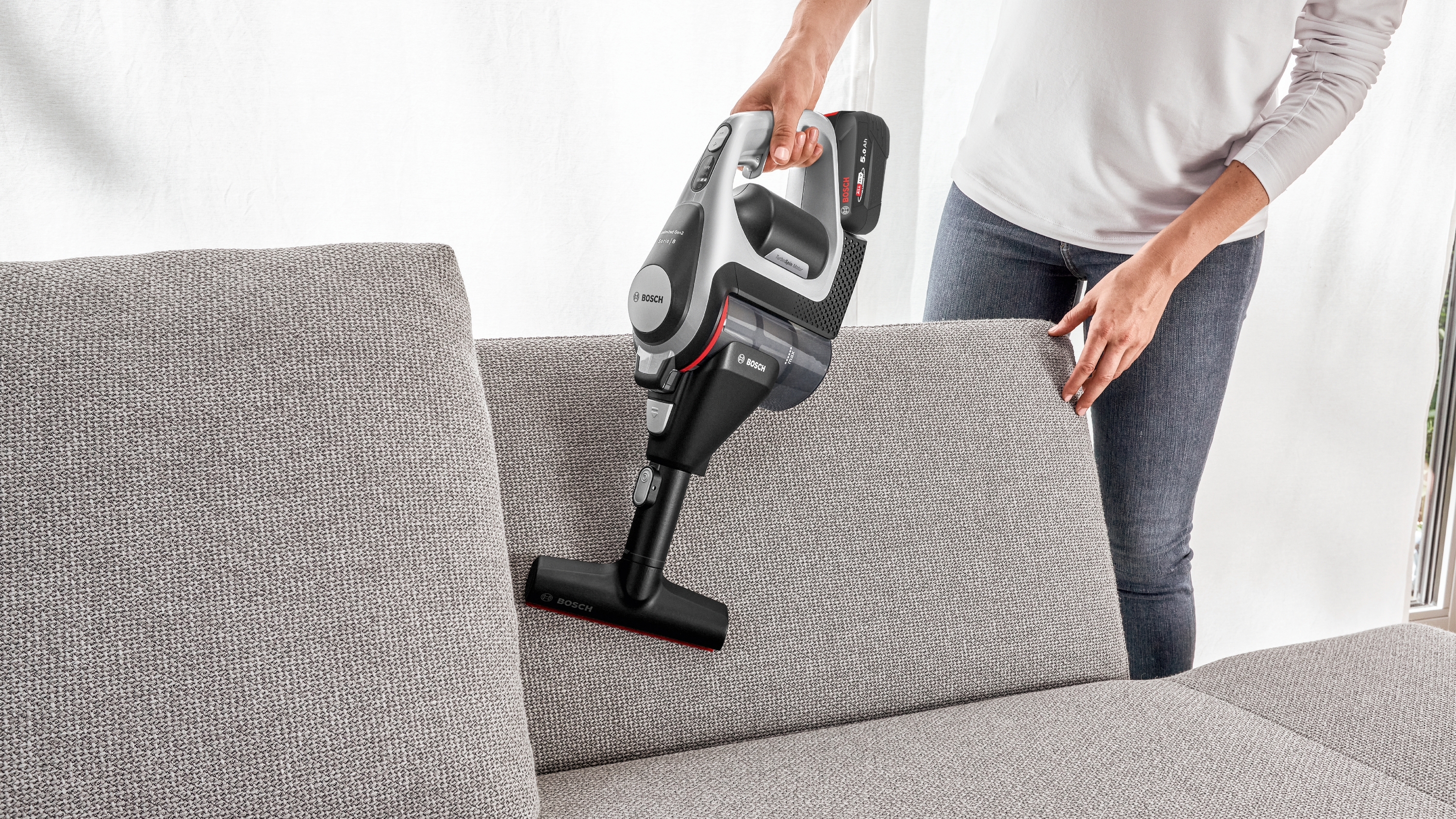 Eine Person, die einen handgehaltenen schwarzen und roten Staubsauger auf einem grauen Sofa benutzt.