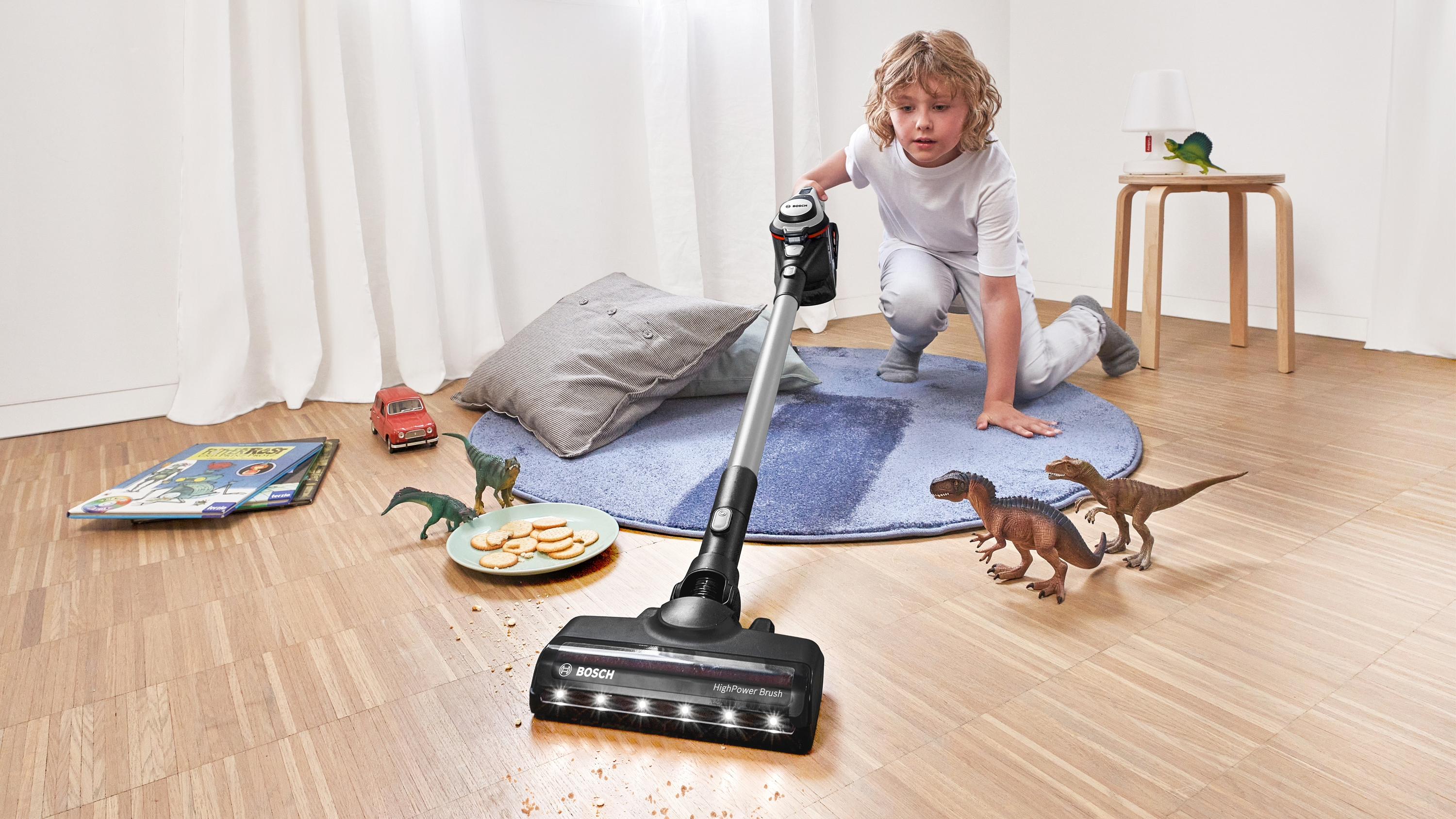 Ein Junge saugt einen Boden mit einem Bosch-Staubsauger, mit Dinosaurierspielzeug und Spielzeugautos.