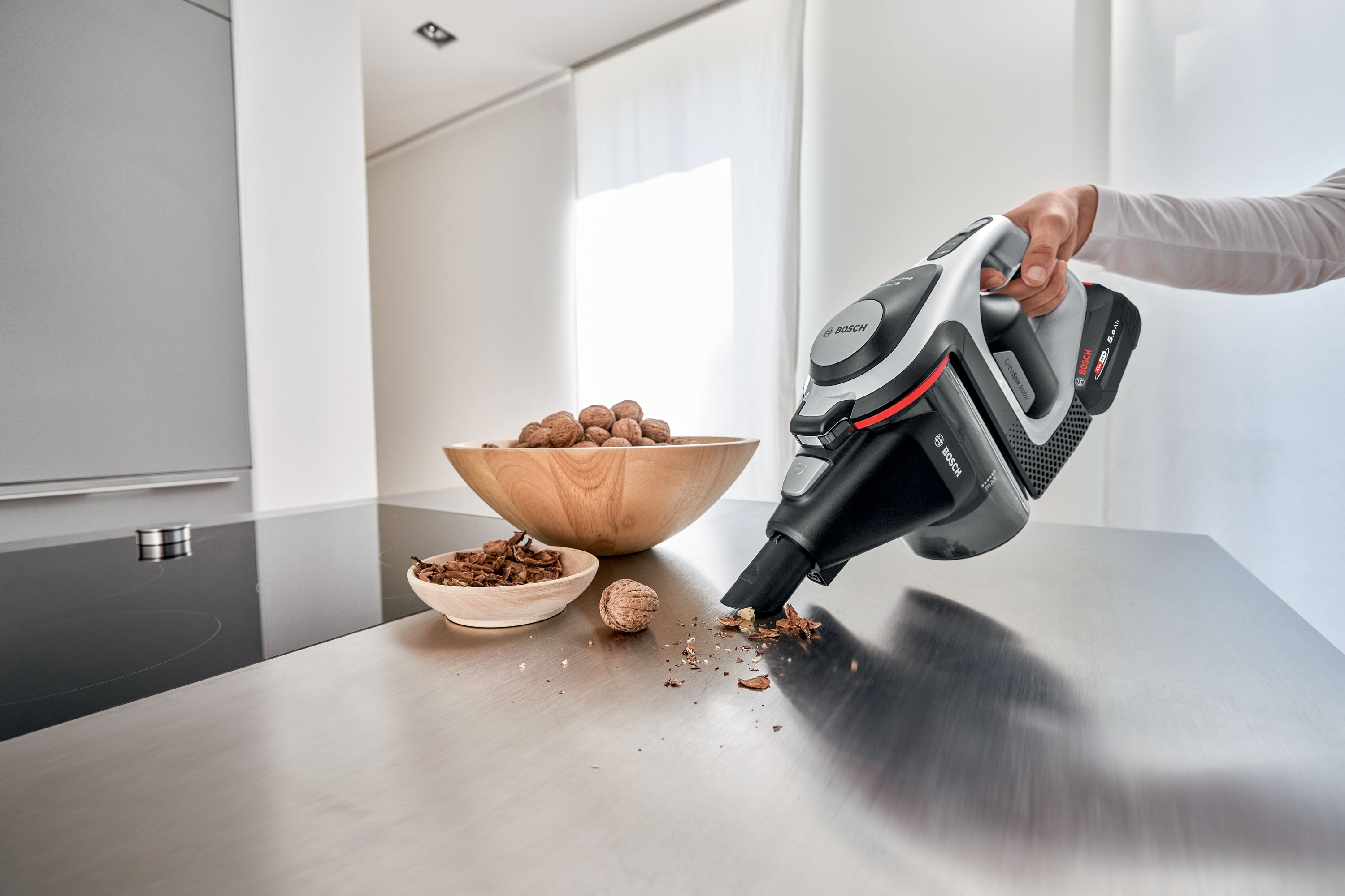 Person saugt mit Bosch-Staubsauger auf Edelstahl-Arbeitsplatte. Schalen mit Nüssen sind da.