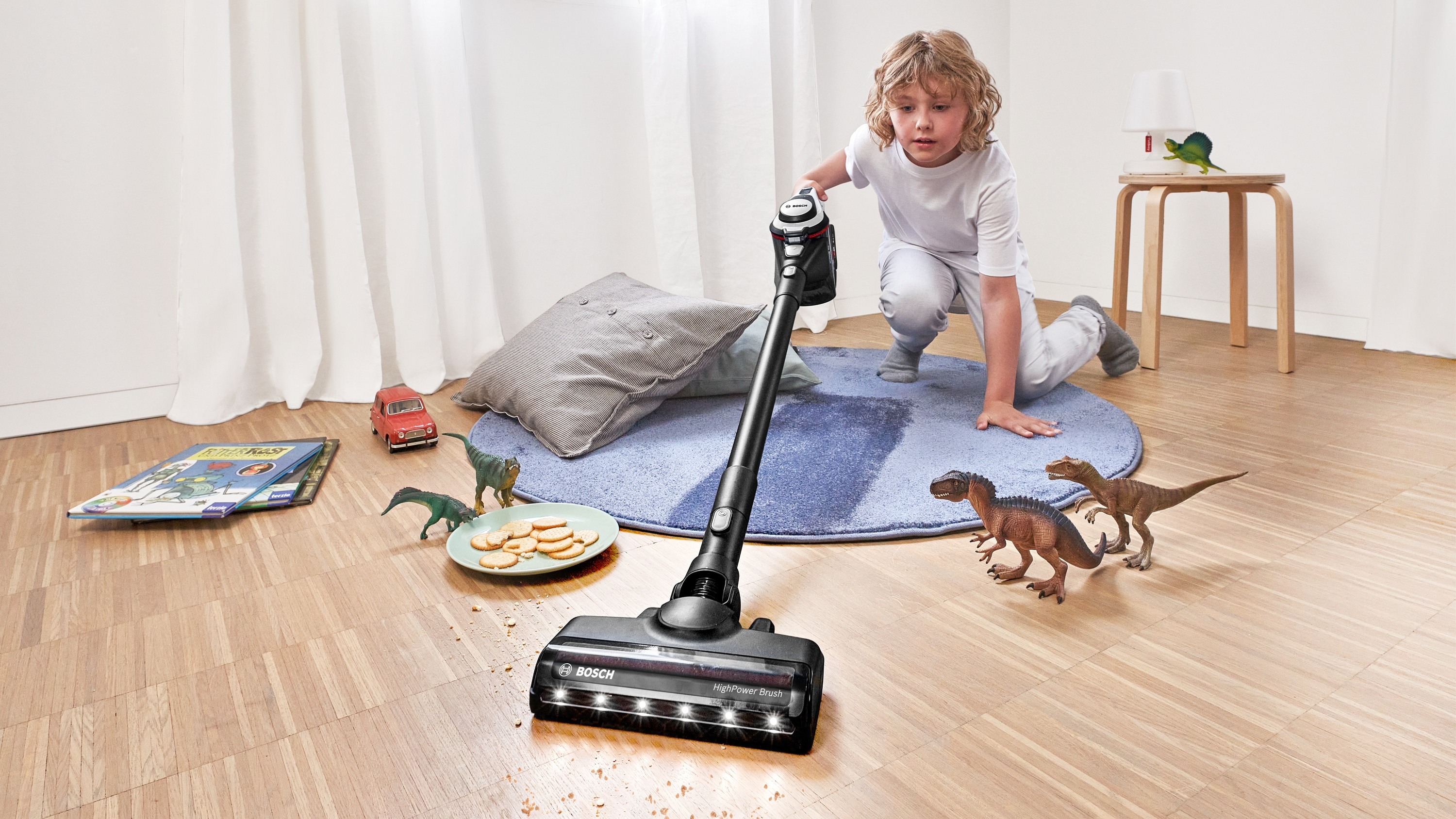 Ein Junge saugt ein Zimmer mit Spielzeug-Dinosauriern, einem Auto und einem Buch auf dem Boden.
