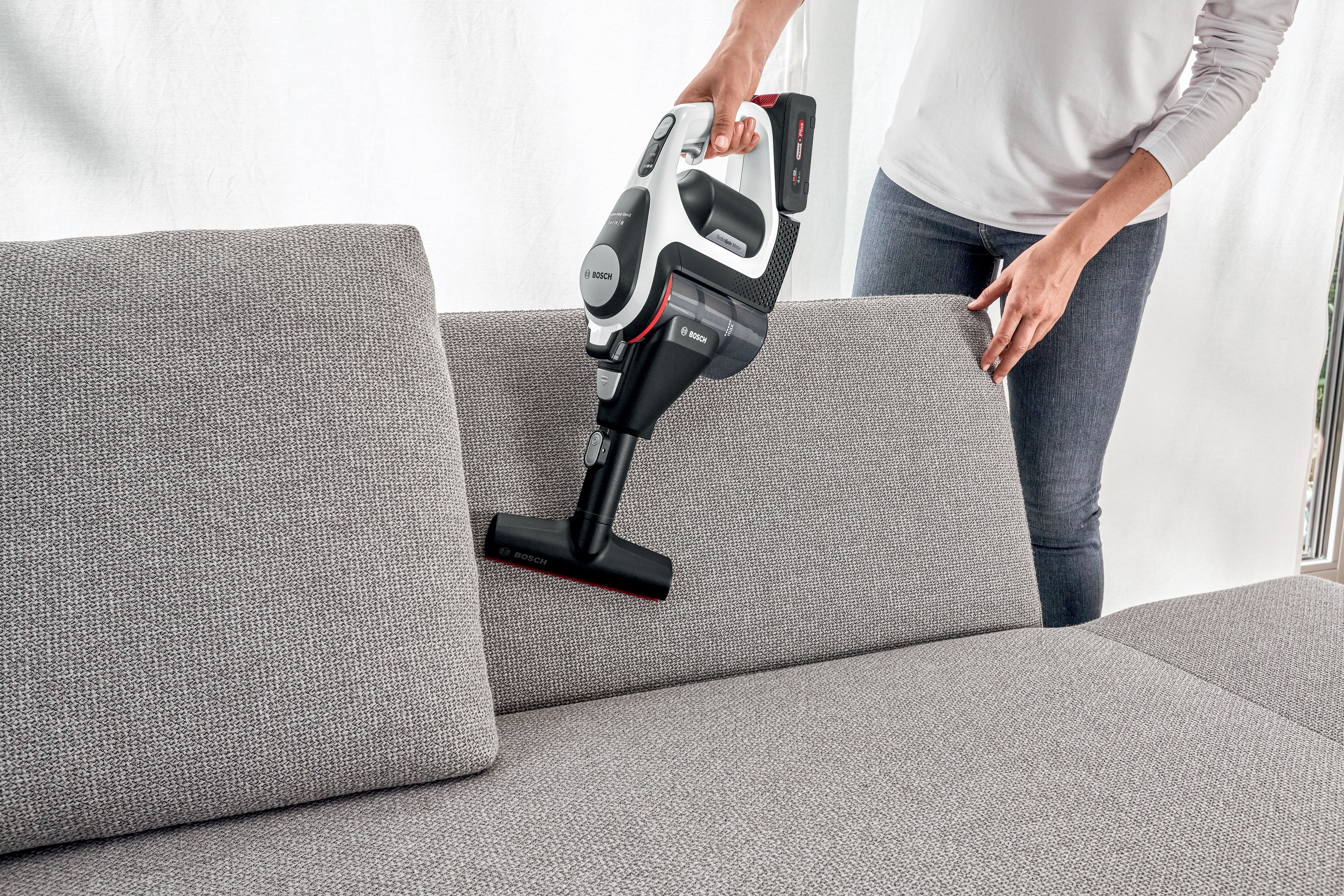 Eine Frau hält einen Staubsauger. Auf der rechten Seite befindet sich ein graues Sofa. Der Hintergrund ist weiß.