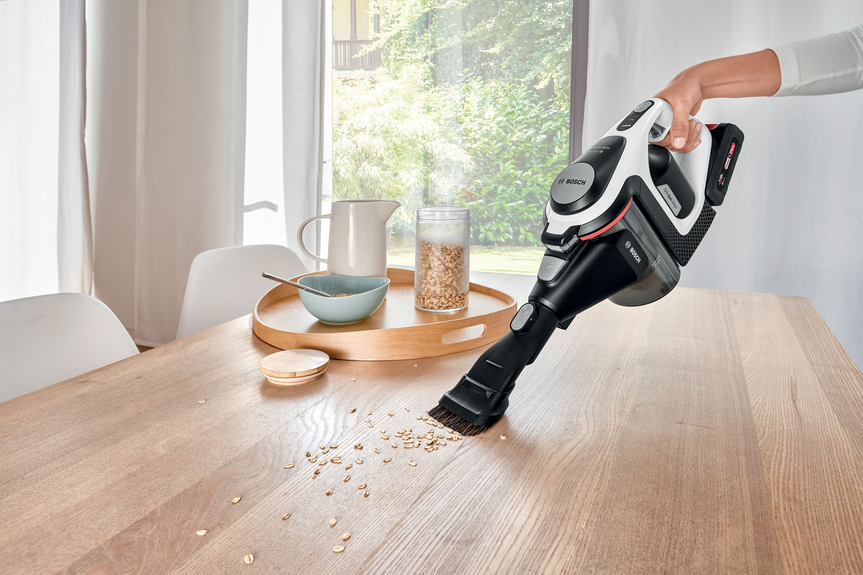 Hand saugt Holztisch ab. Verstreute Haferflocken sind auf dem Tisch sichtbar.