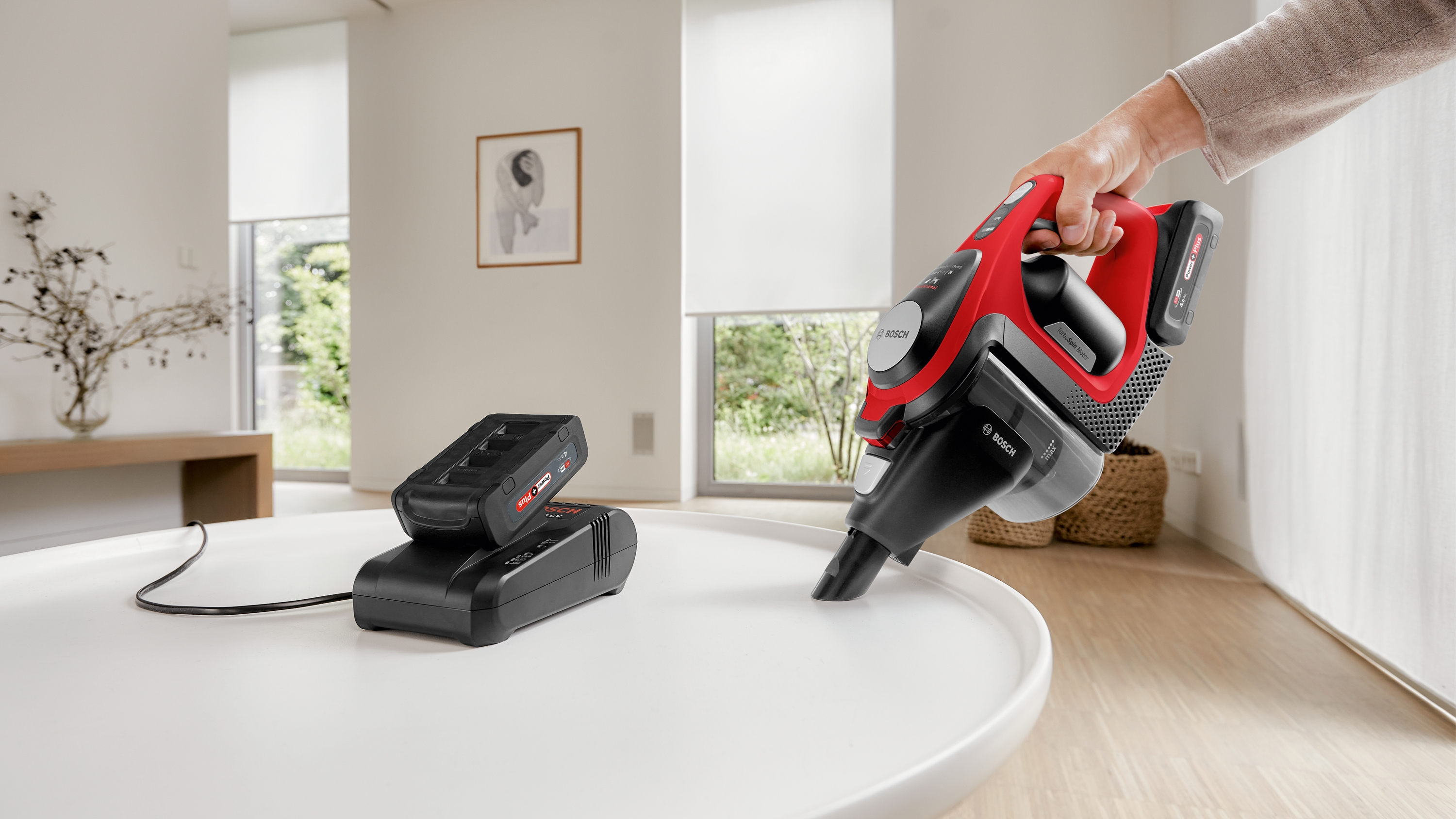 Ein rot-schwarzer Staubsauger reinigt einen weißen Tisch. Eine Ladestation ist in der Nähe.
