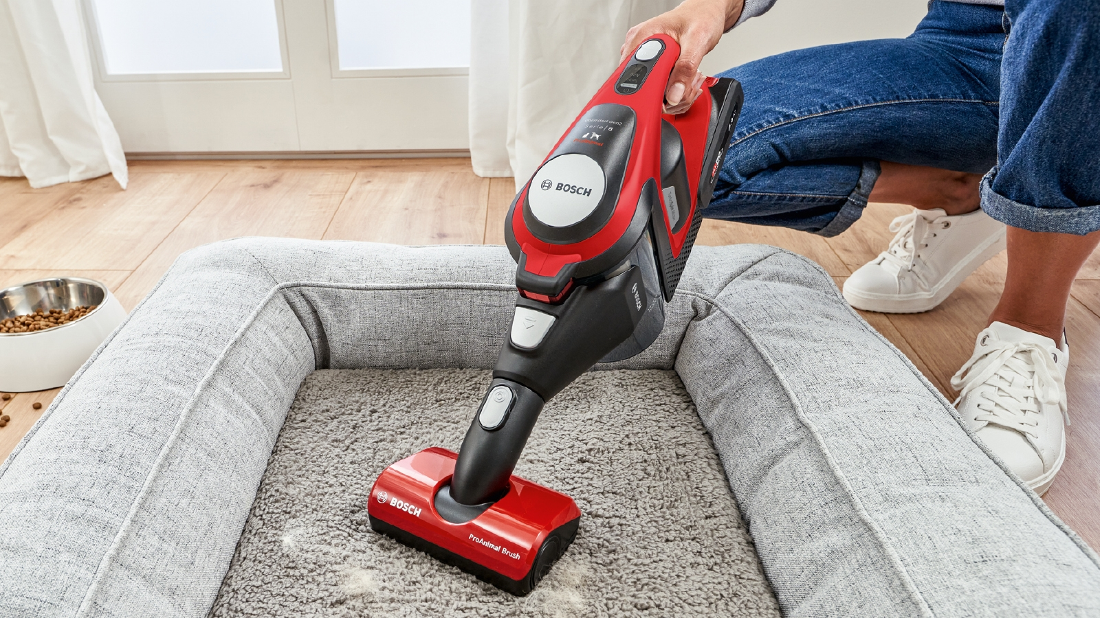 Eine Person benutzt einen rot-schwarzen Akku-Staubsauger auf einem Hundebett.
