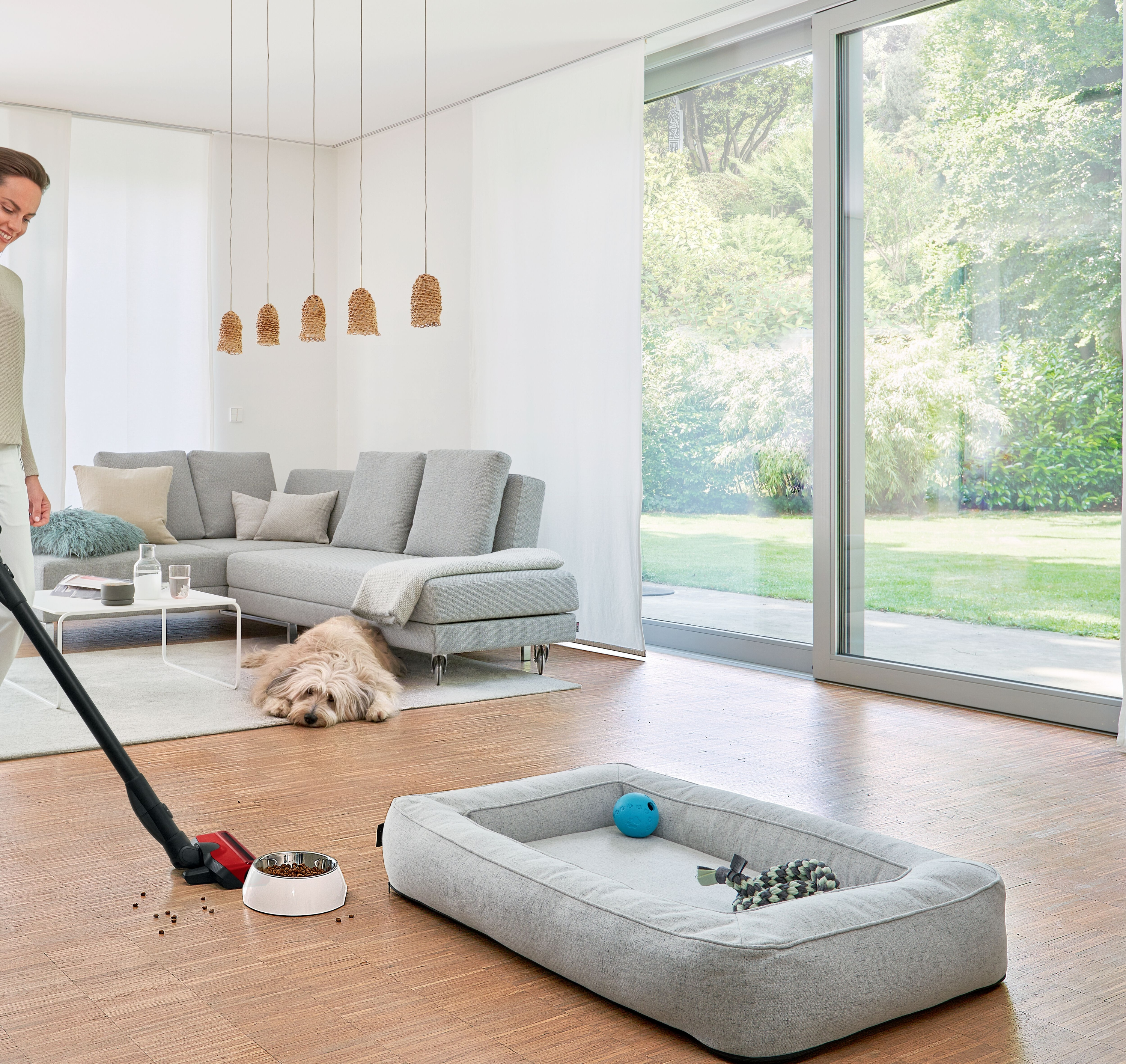 Rauminnenraum mit einer Frau, die staubsaugt. Ein Hund ruht neben einem Hundebett. Sofa und Fenster im Blick.