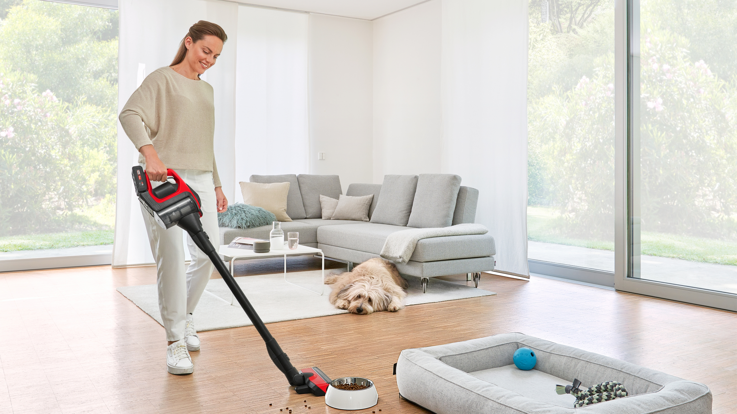 Frau saugt Holzboden, Hund ruht. Sofa, Fenster und Hundebett im Hintergrund.