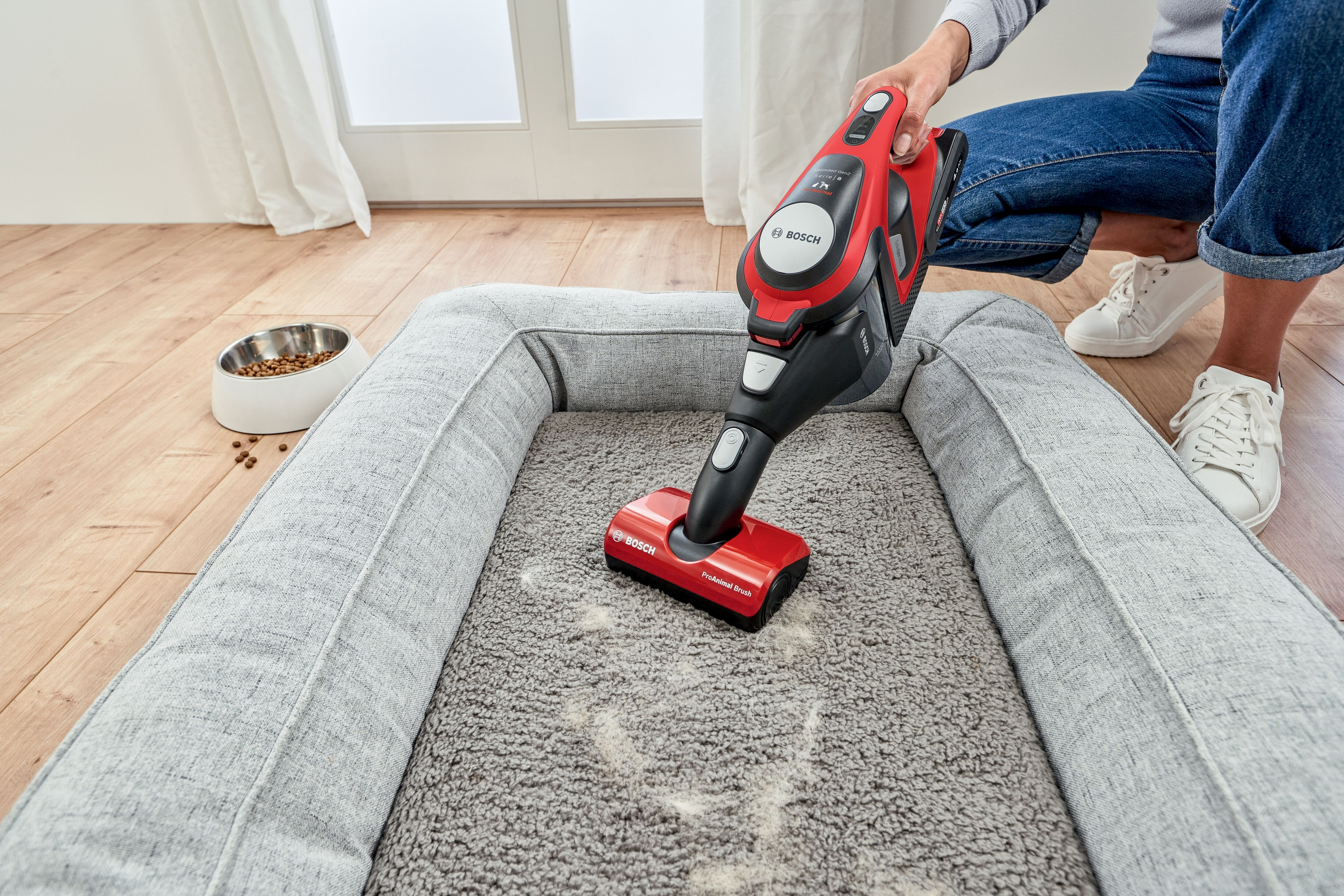 Eine Person, die mit einem rot-schwarzen Bosch-Staubsauger ein Hundebett absaugt; eine Futterschüssel in der Nähe.