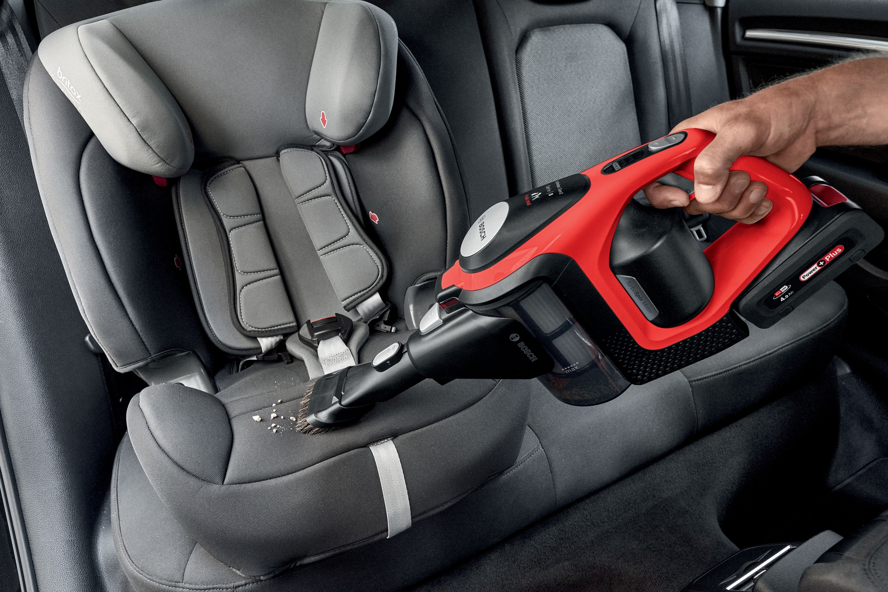Eine Hand, die einen rot-schwarzen Staubsauger in einem Auto benutzt. Kindersitz im Hintergrund.