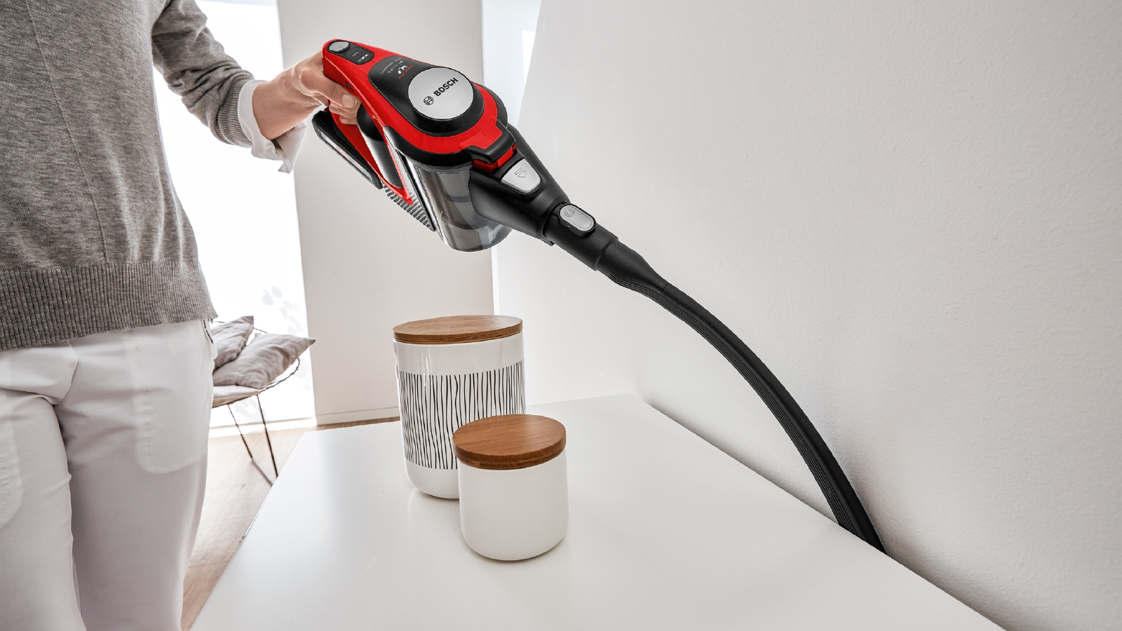 Eine Hand, die einen rot-schwarzen kabellosen Staubsauger hält und eine weiße Oberfläche reinigt.