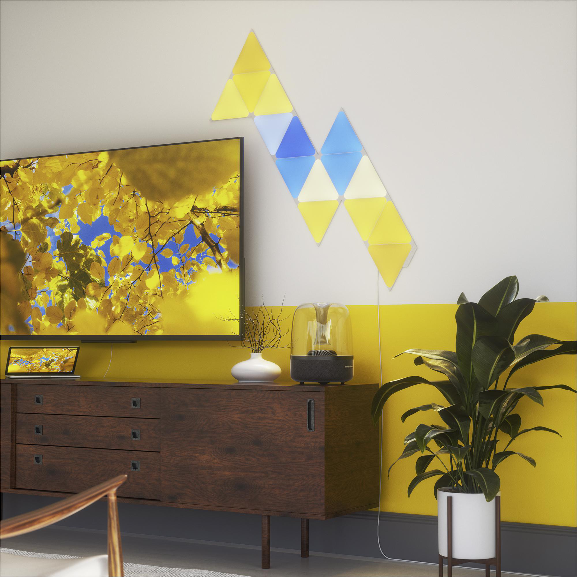 Raum mit Fernseher, Laptop und Pflanze. Wandkunst aus gelben und blauen Dreiecken.