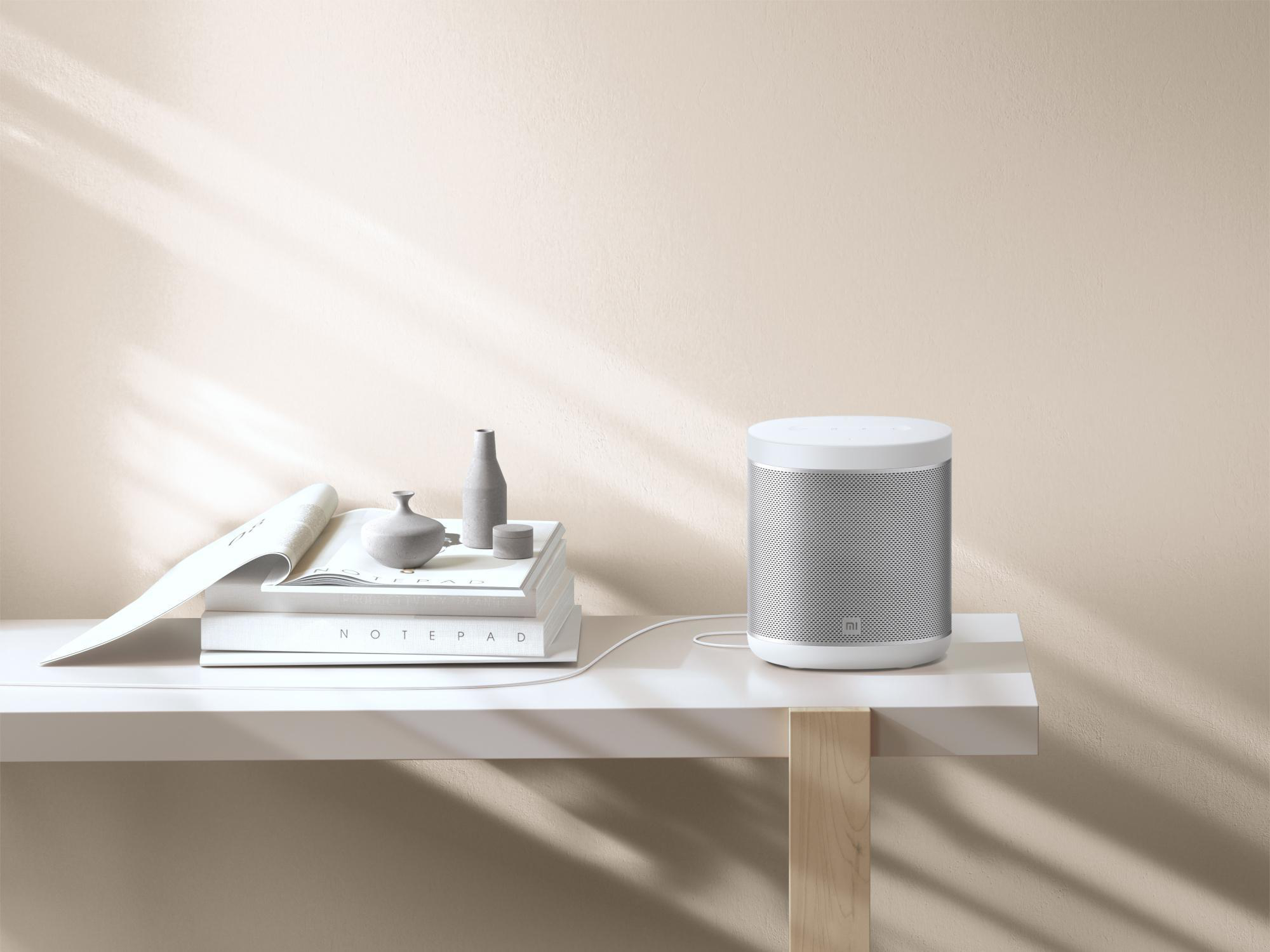 Weißer Smart Speaker auf einem weißen Regal. Bücher und Dekoration stehen daneben.