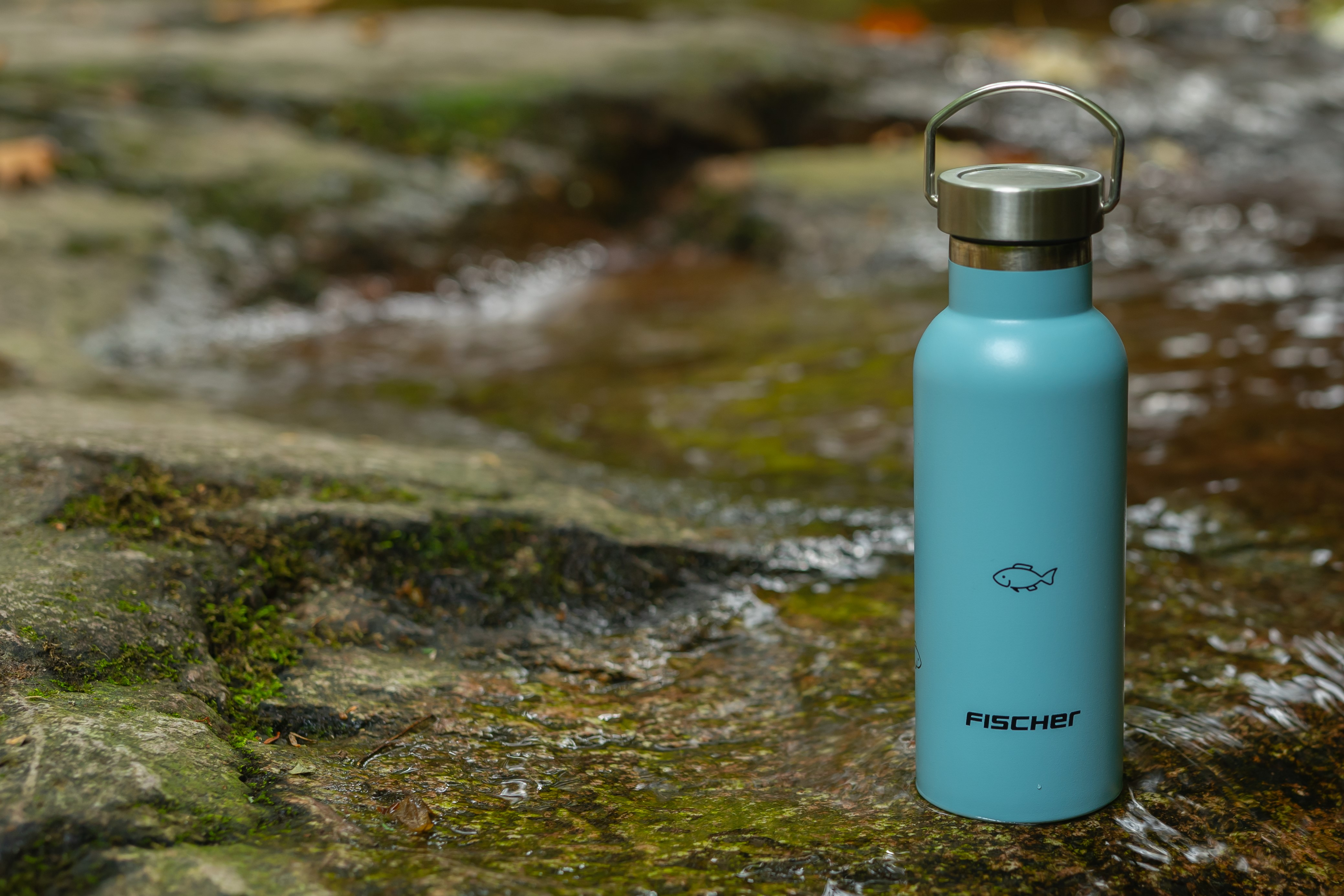 Blaue Wasserflasche mit Fischlogo, auf Felsen in der Nähe von Wasser mit verschwommenem Hintergrund.