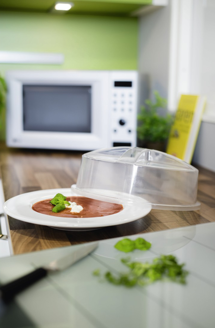 Een wit bord met soep en een plastic deksel. Een magnetron, mes en plant staan op de achtergrond.