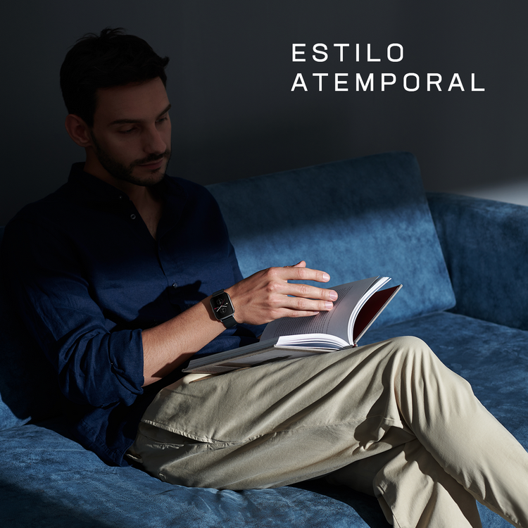 Un hombre leyendo un libro en un sofá azul, con reloj y pantalones color caqui. Texto: Estilo atemporal.