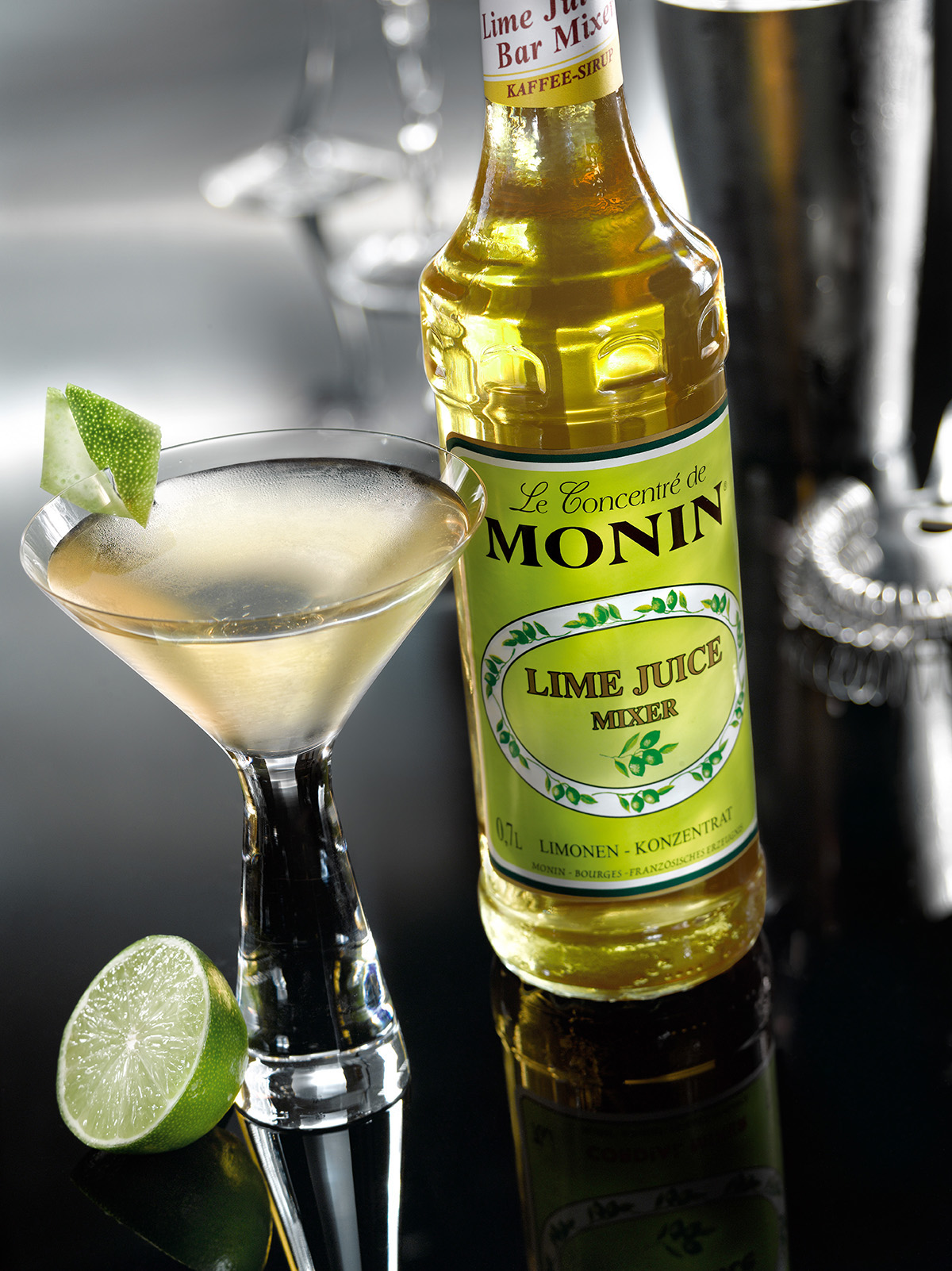 Ein Cocktailglas mit Getränk, Limette und einer Flasche Monin Limettensaft.