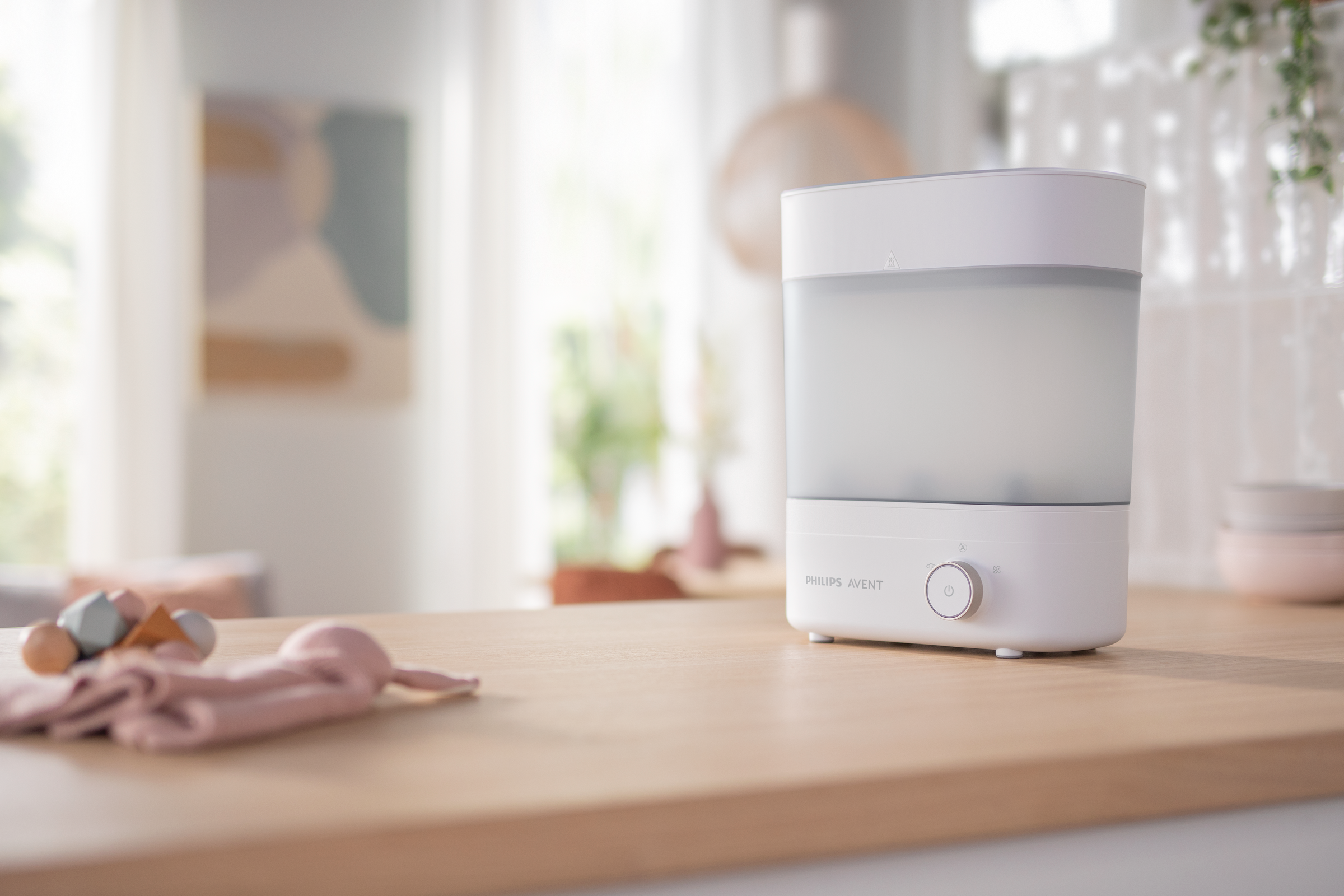 Sterilizzatore per biberon bianco su superficie in legno, con giocattoli per bambini sullo sfondo.