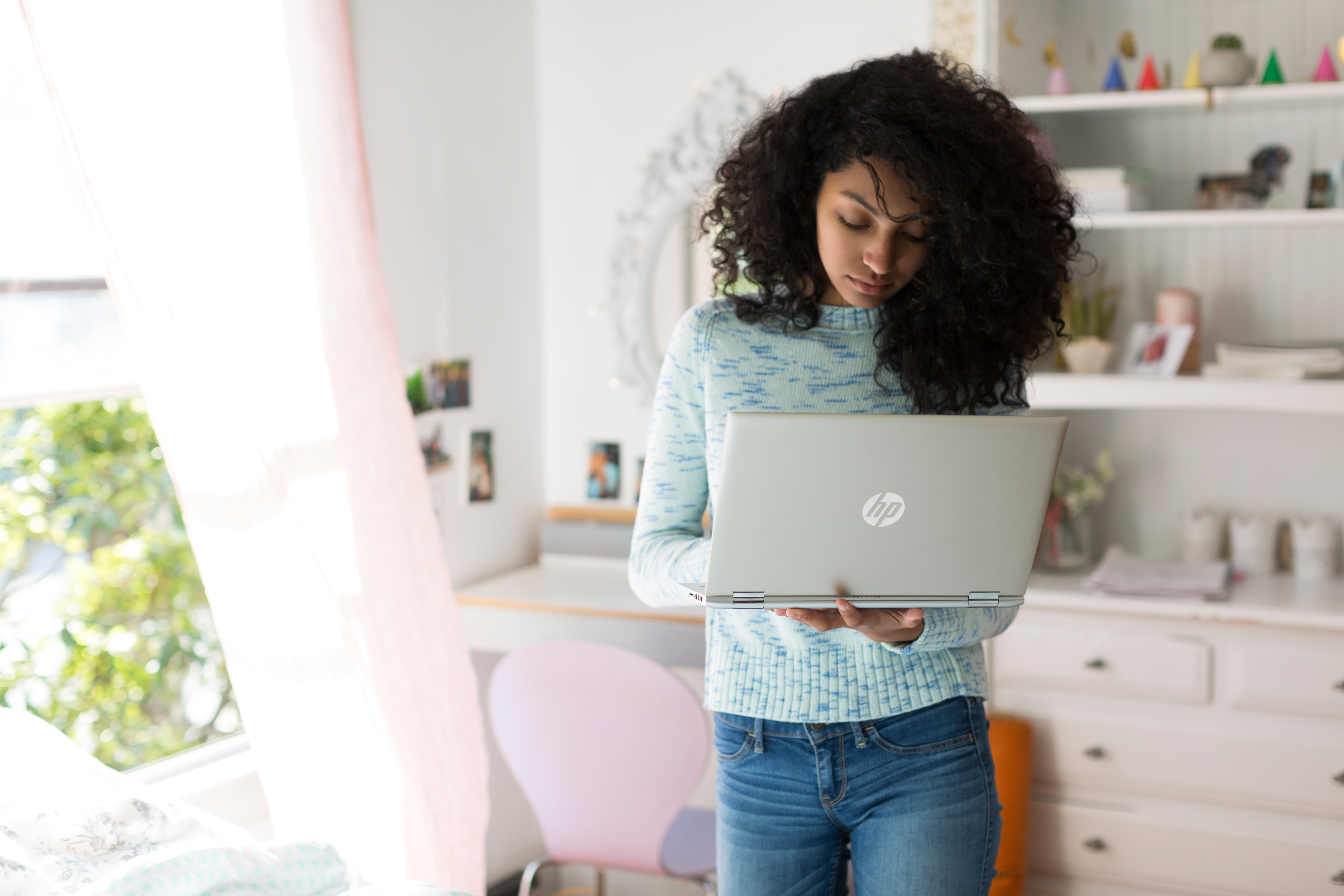 Eine junge Frau mit lockigem Haar steht in einem Zimmer und hält einen Laptop.