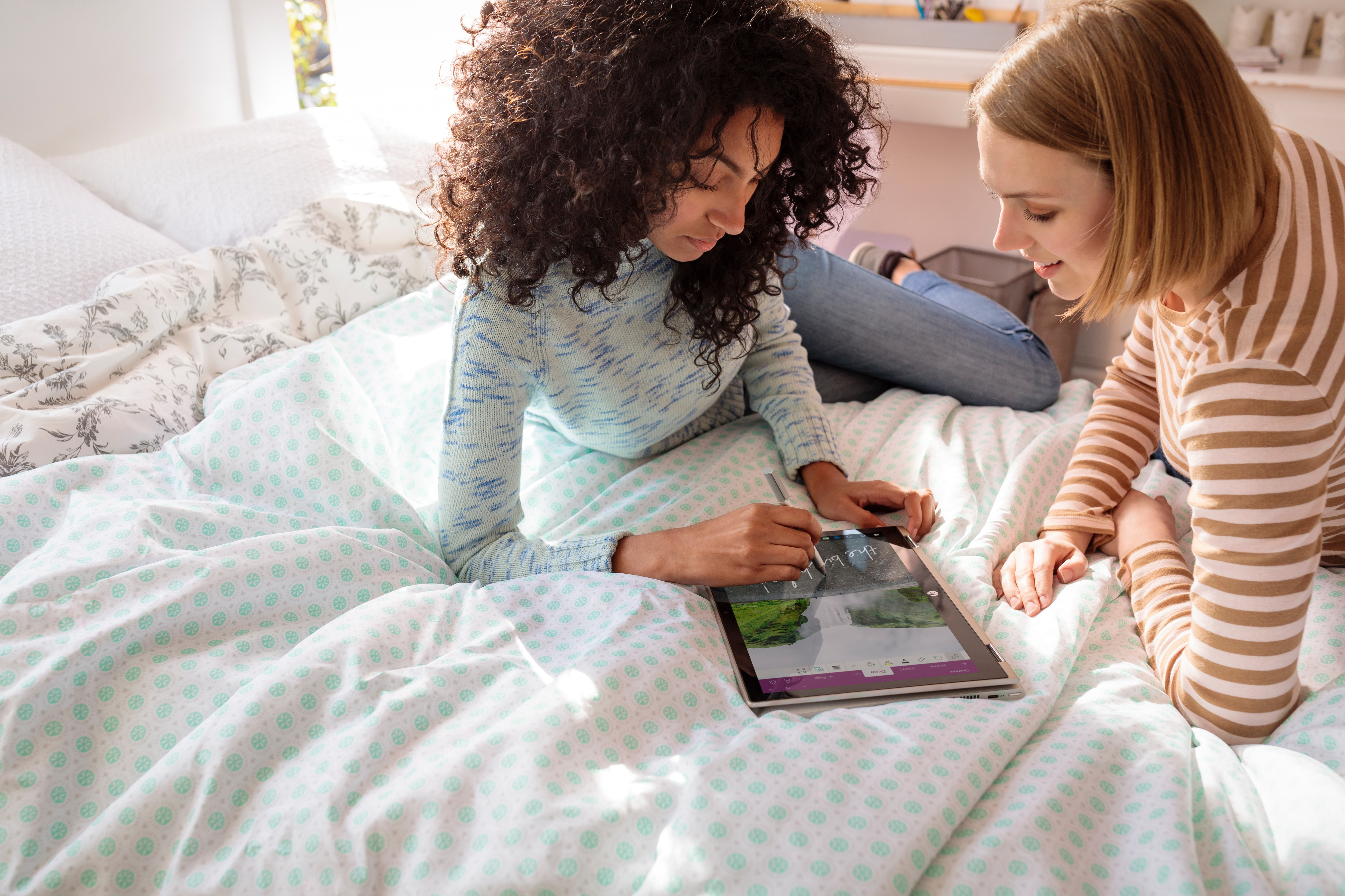 Zwei junge Frauen liegen auf einem gemusterten Bett und betrachten ein Tablet.