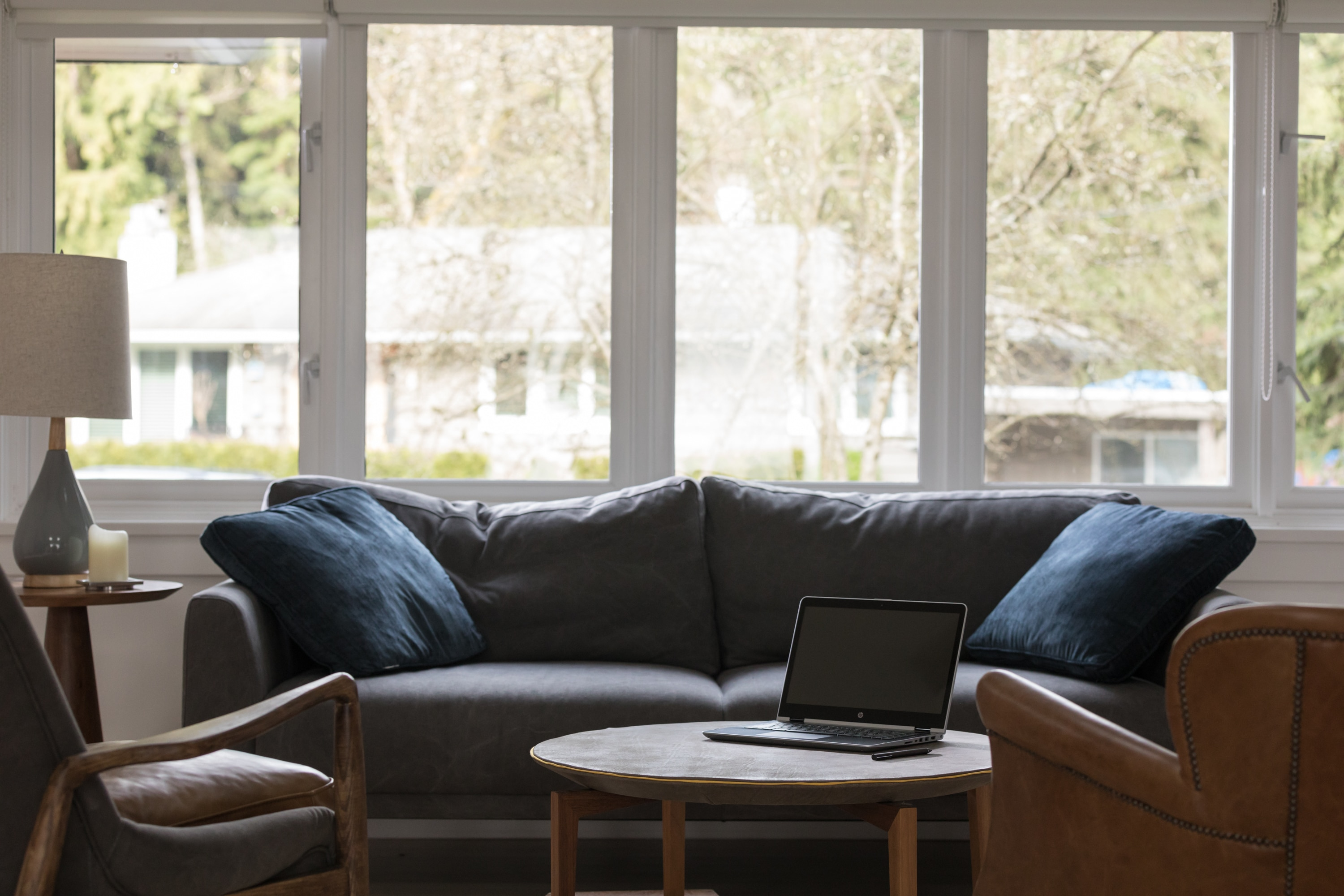 Ein Wohnzimmer mit grauem Sofa, blauen Kissen, Laptop auf dem Couchtisch und großen Fenstern.