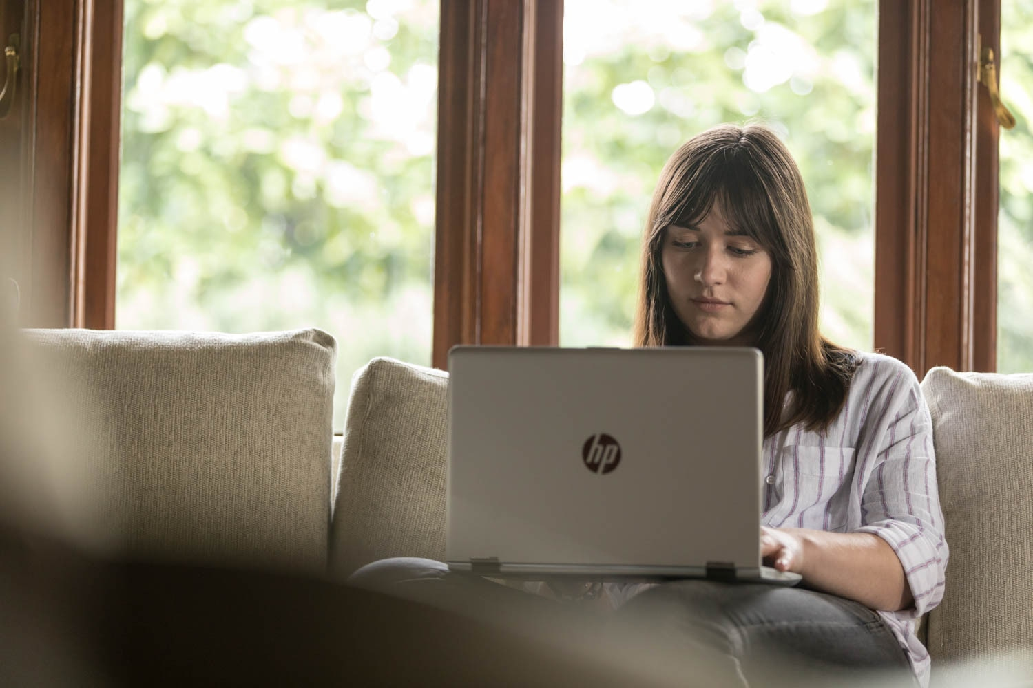 Eine junge Frau sitzt vor einem Fenster auf einem Sofa und arbeitet an einem HP Laptop.