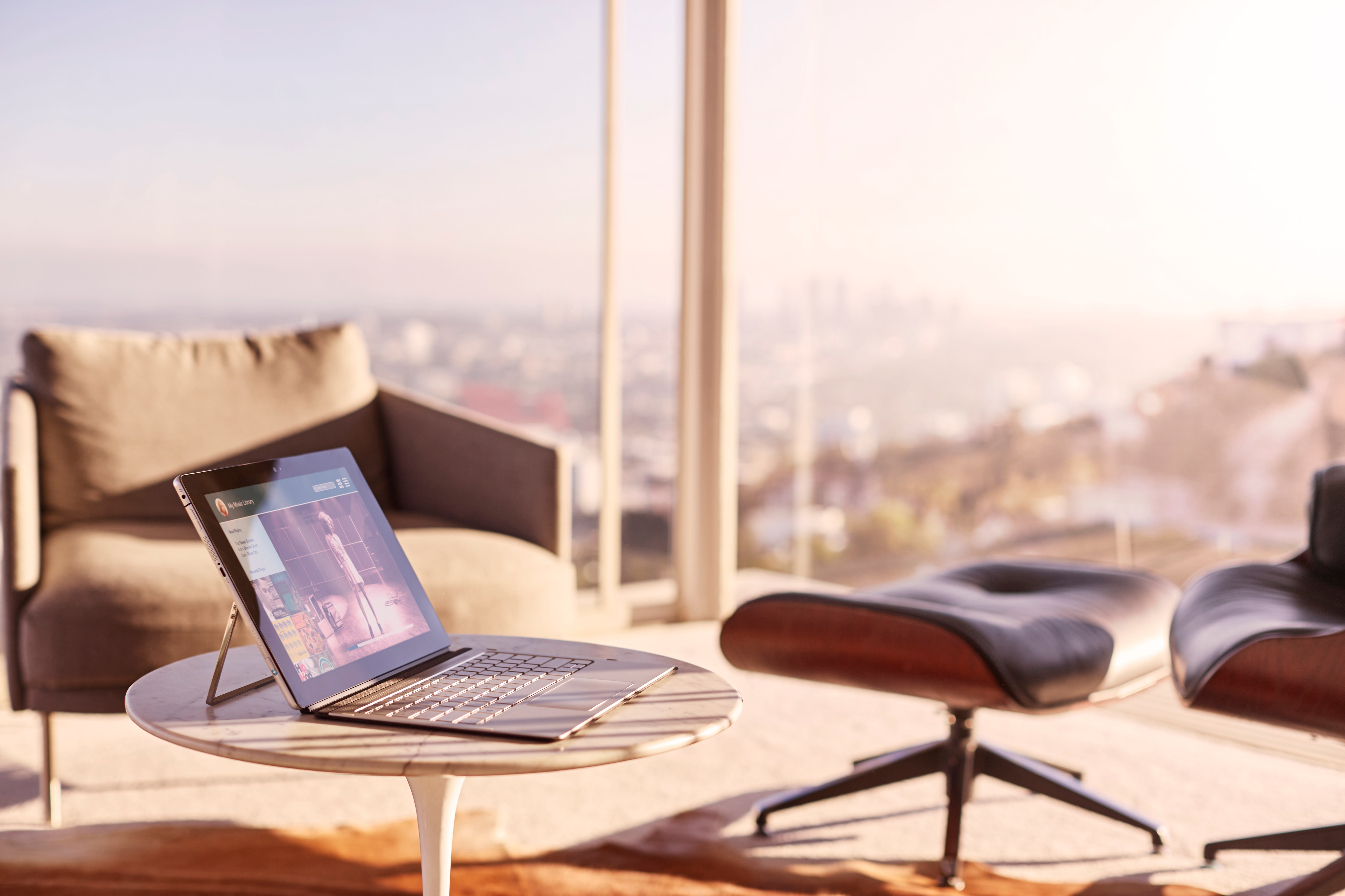 Ein Laptop steht auf einem runden Tisch in einem hellen Raum mit Sesseln und weitem Stadtblick.