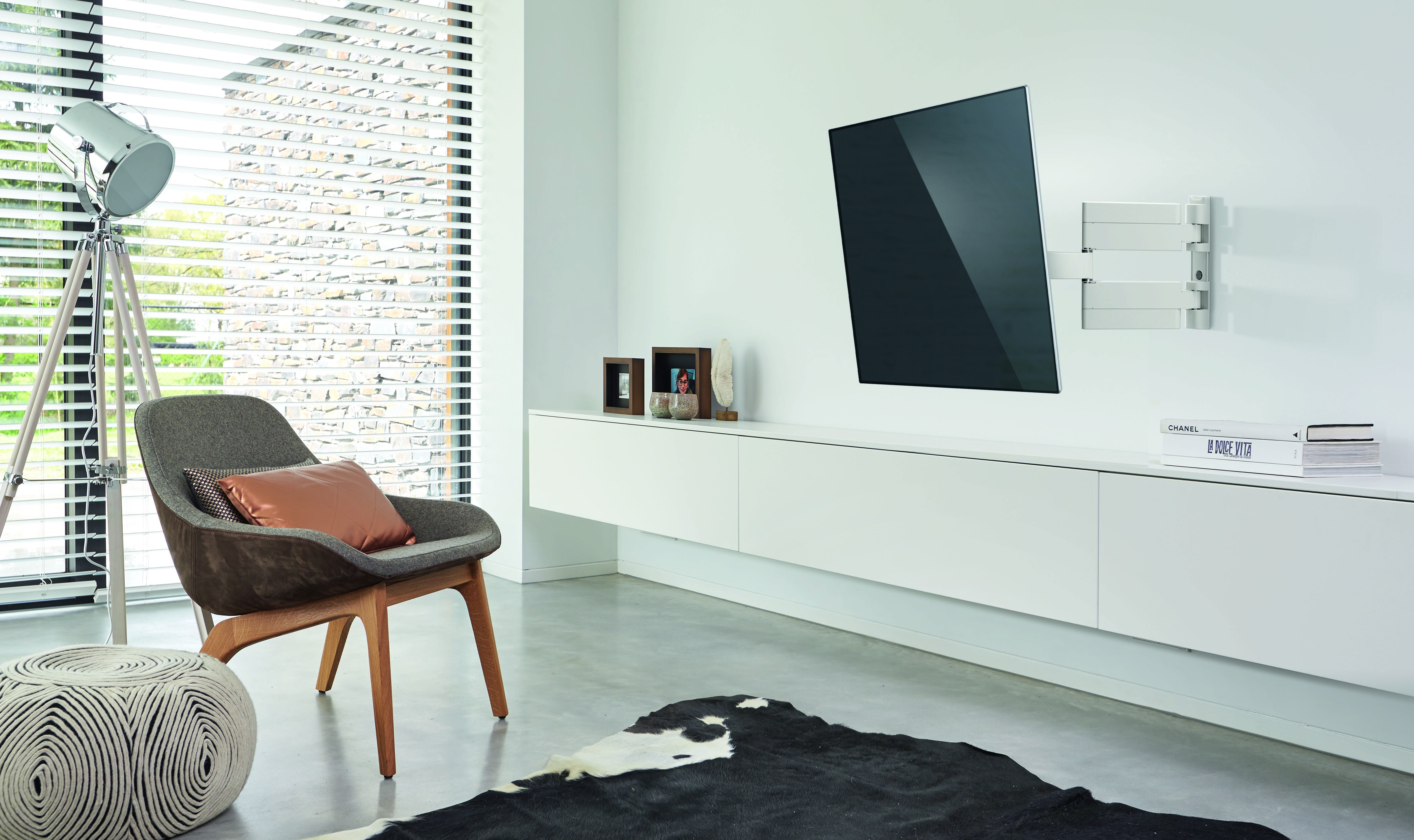 Modernes Wohnzimmer mit TV an der Wand, grauem Stuhl und weißen Möbeln. Ein Kuhfellteppich liegt auf dem Boden.