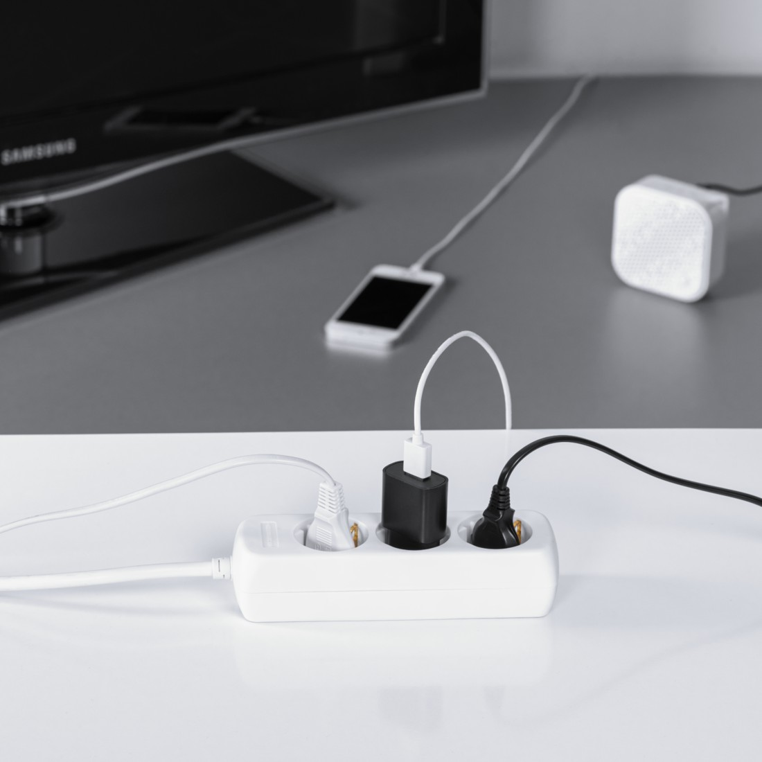 Ein weißer Mehrfachstecker mit drei angeschlossenen Geräten und einem Fernseher im Hintergrund.