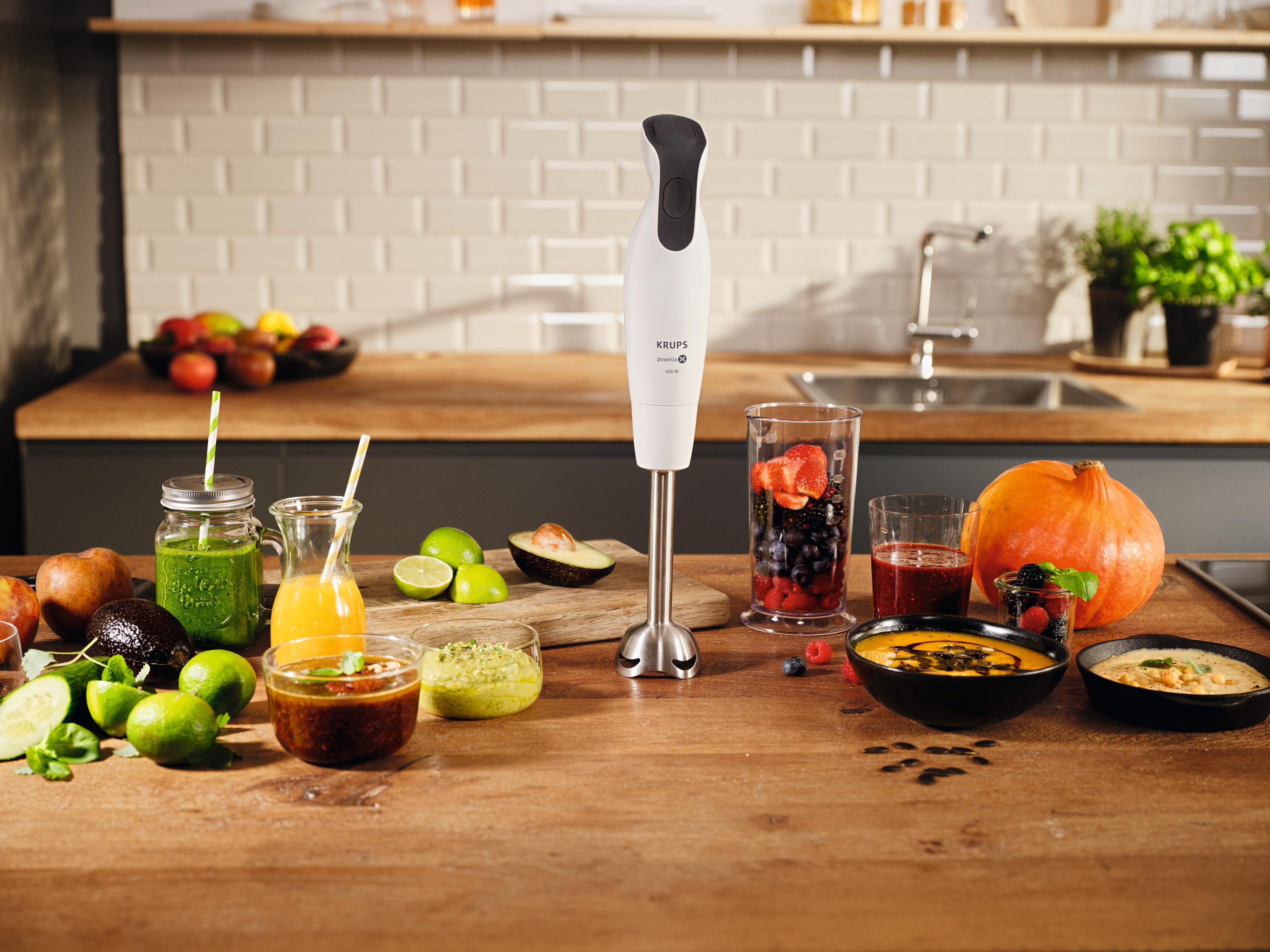 Holztisch mit Mixer, Lebensmitteln und Zutaten. Küchenhintergrund mit Personen.