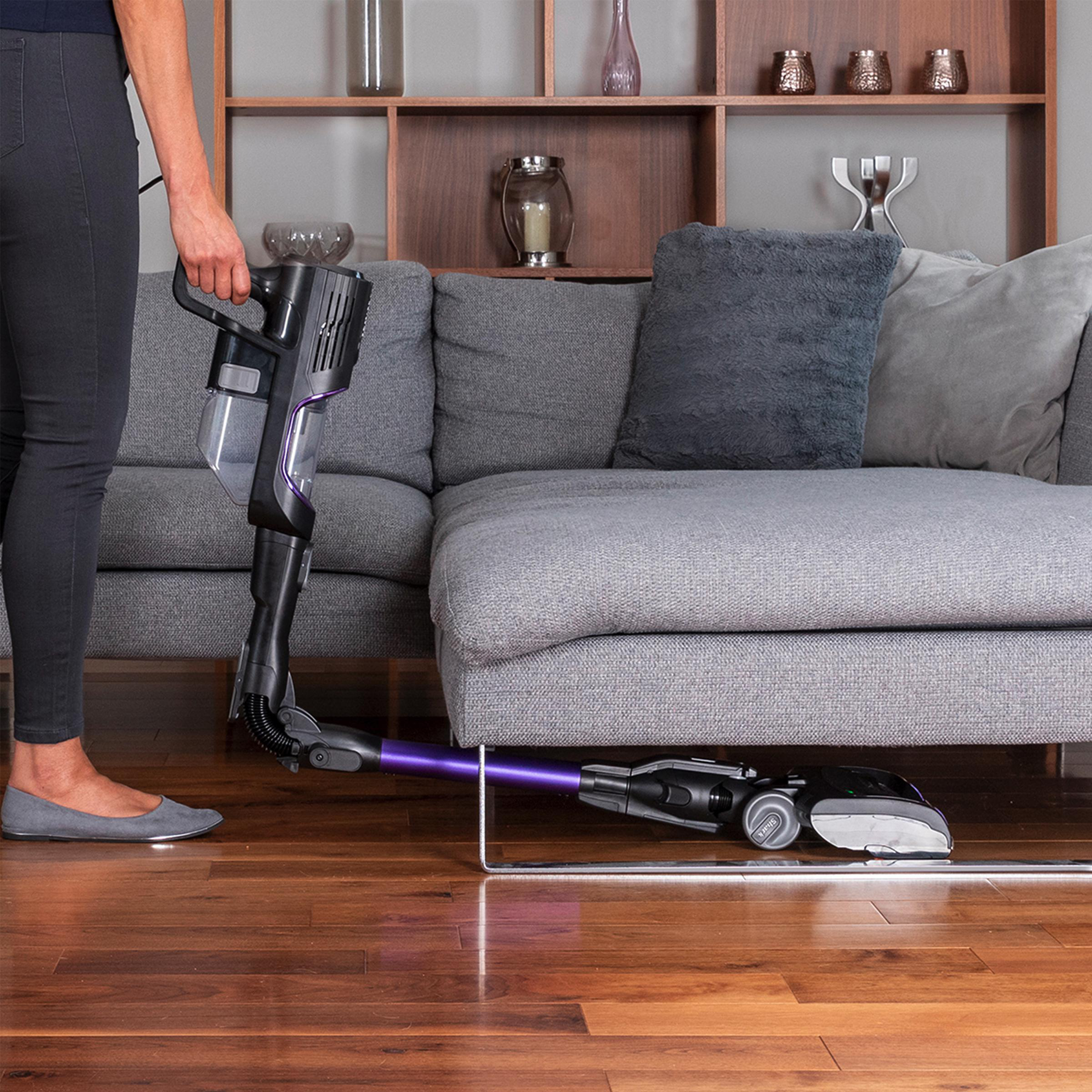 Person hält Staubsauger, reinigt unter einem grauen Sofa. Holzboden.