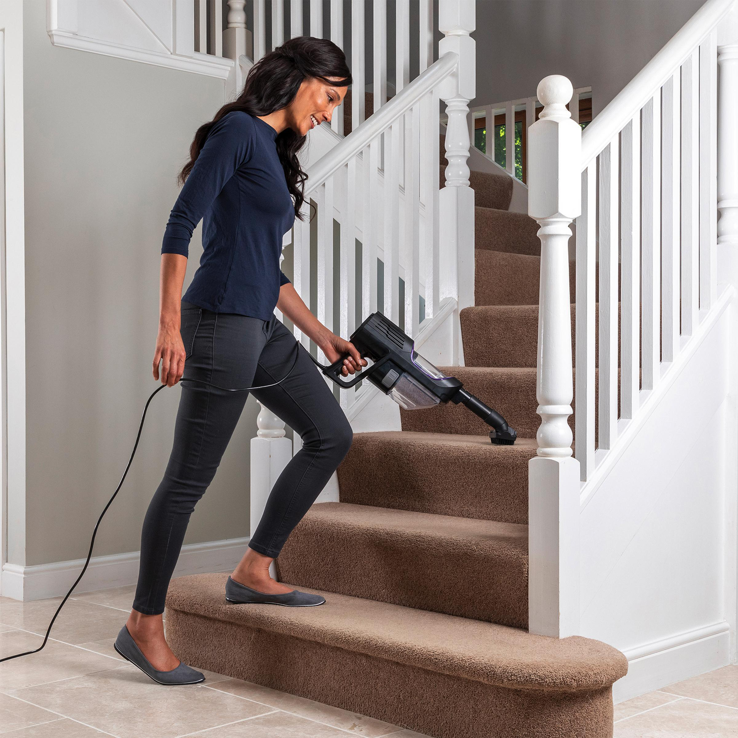 Eine Frau, die mit einem schwarzen Shark-Staubsauger Treppen saugt. Sie trägt dunkle Kleidung. Die Treppe ist mit Teppich ausgelegt.