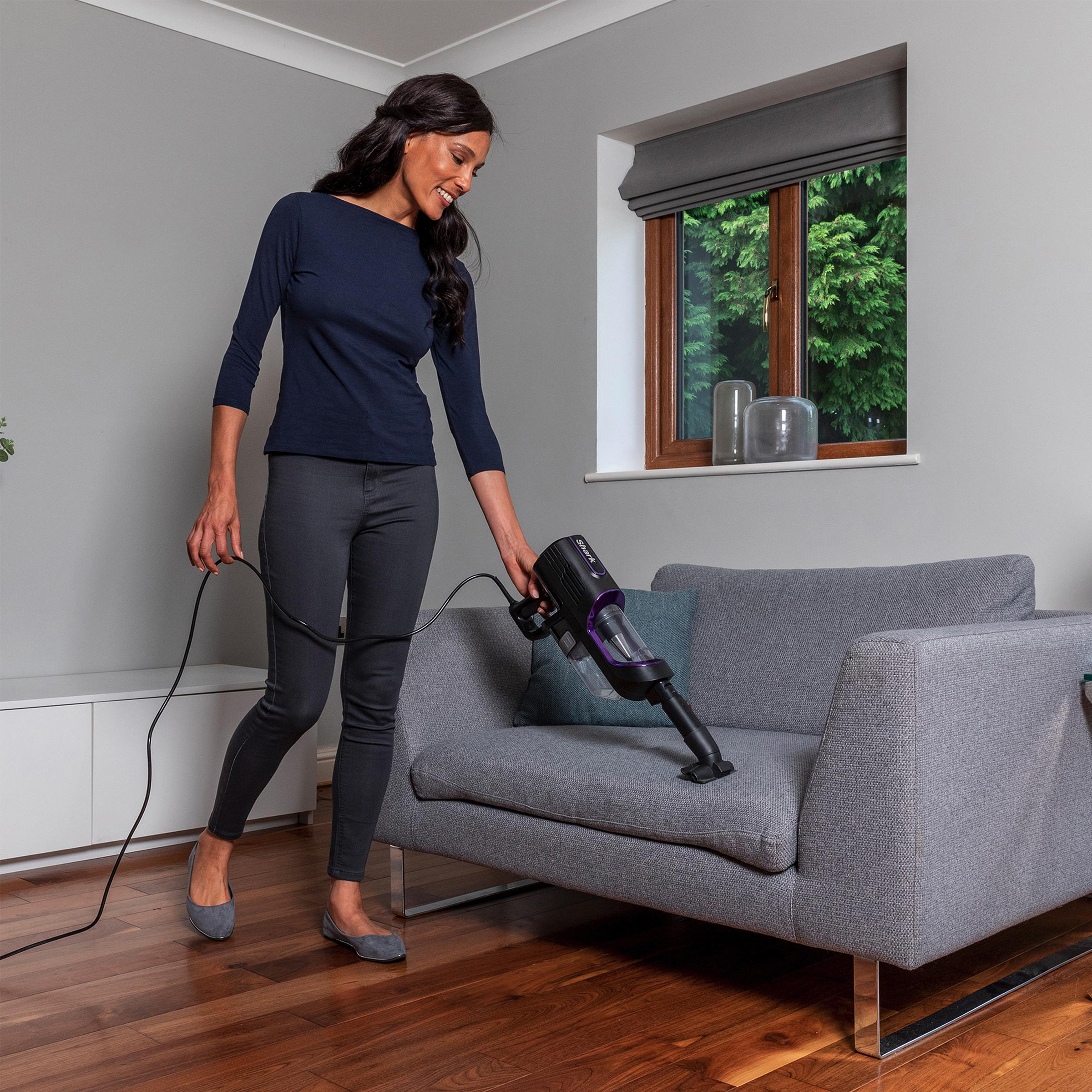 Frau, die mit einem schwarz-violetten Handstaubsauger ein graues Sofa absaugt.