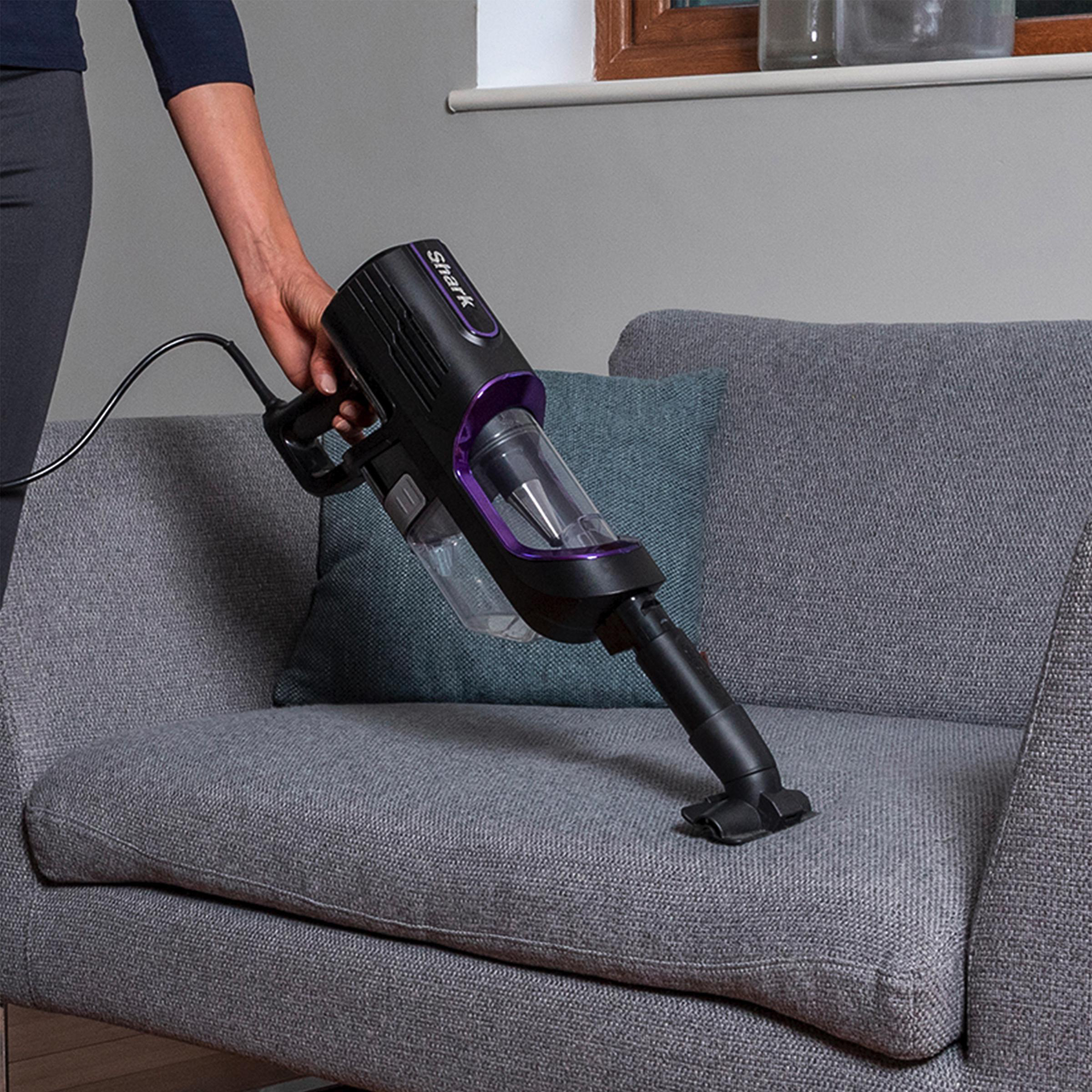 Eine Frau benutzt einen kabellosen Staubsauger in Schwarz und Lila auf einem grauen Sofa.