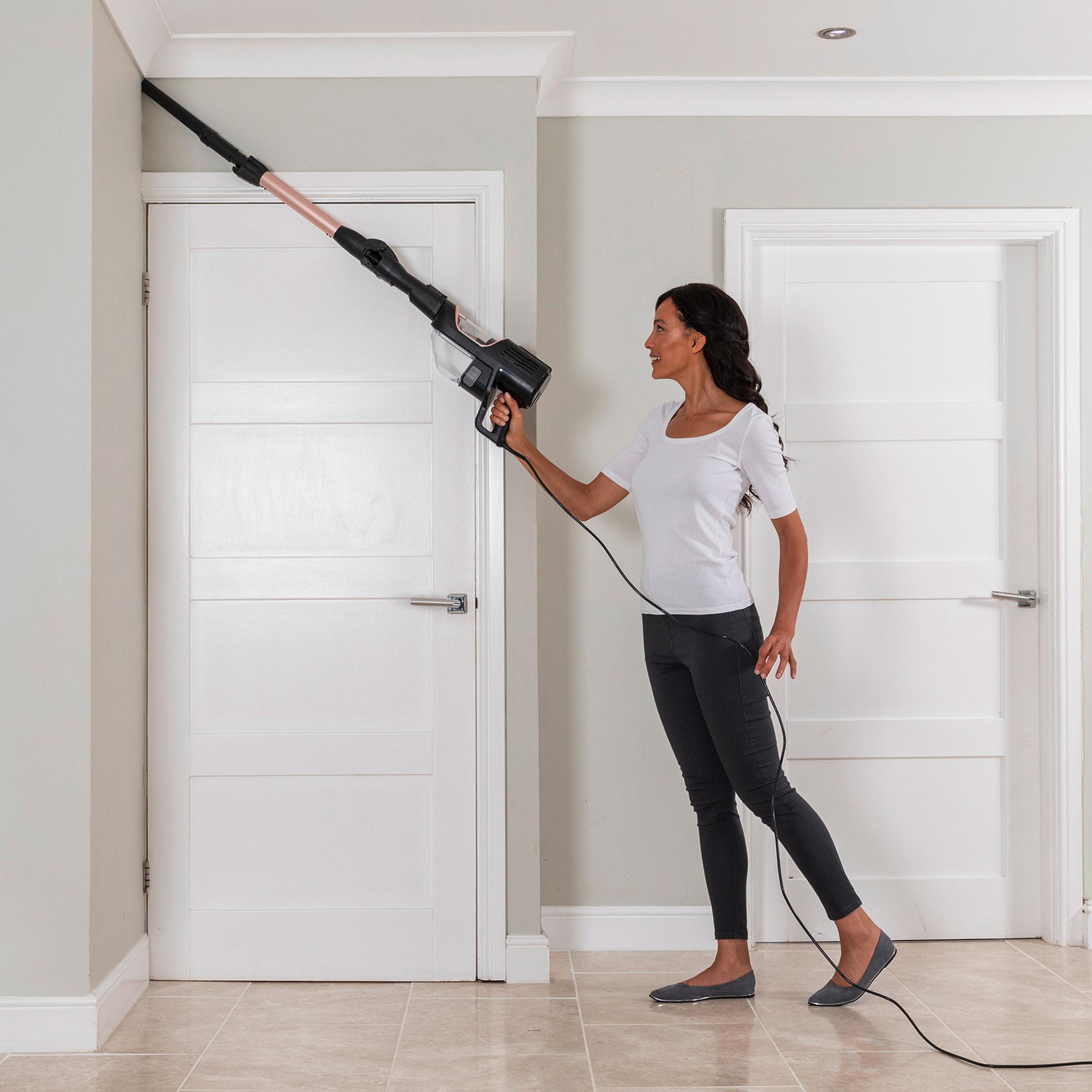 Eine Frau saugt mit einem aufrechten Staubsauger eine Wand ab. Es gibt zwei Türen, eine geschlossen.