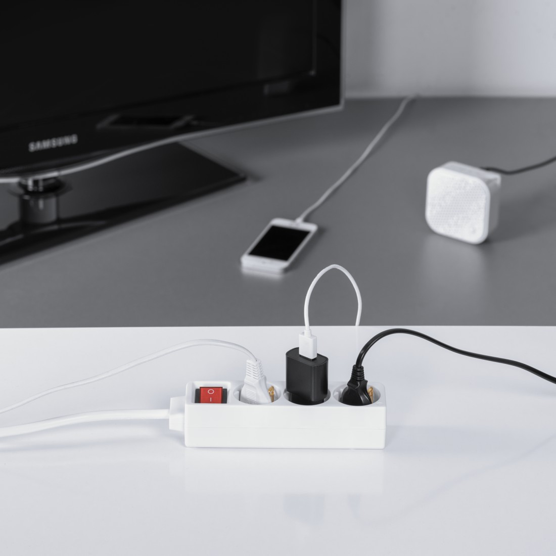 Eine weiße Steckdosenleiste mit rotem Schalter ist an Geräte angeschlossen. Ein Fernseher und ein Handy sind sichtbar.