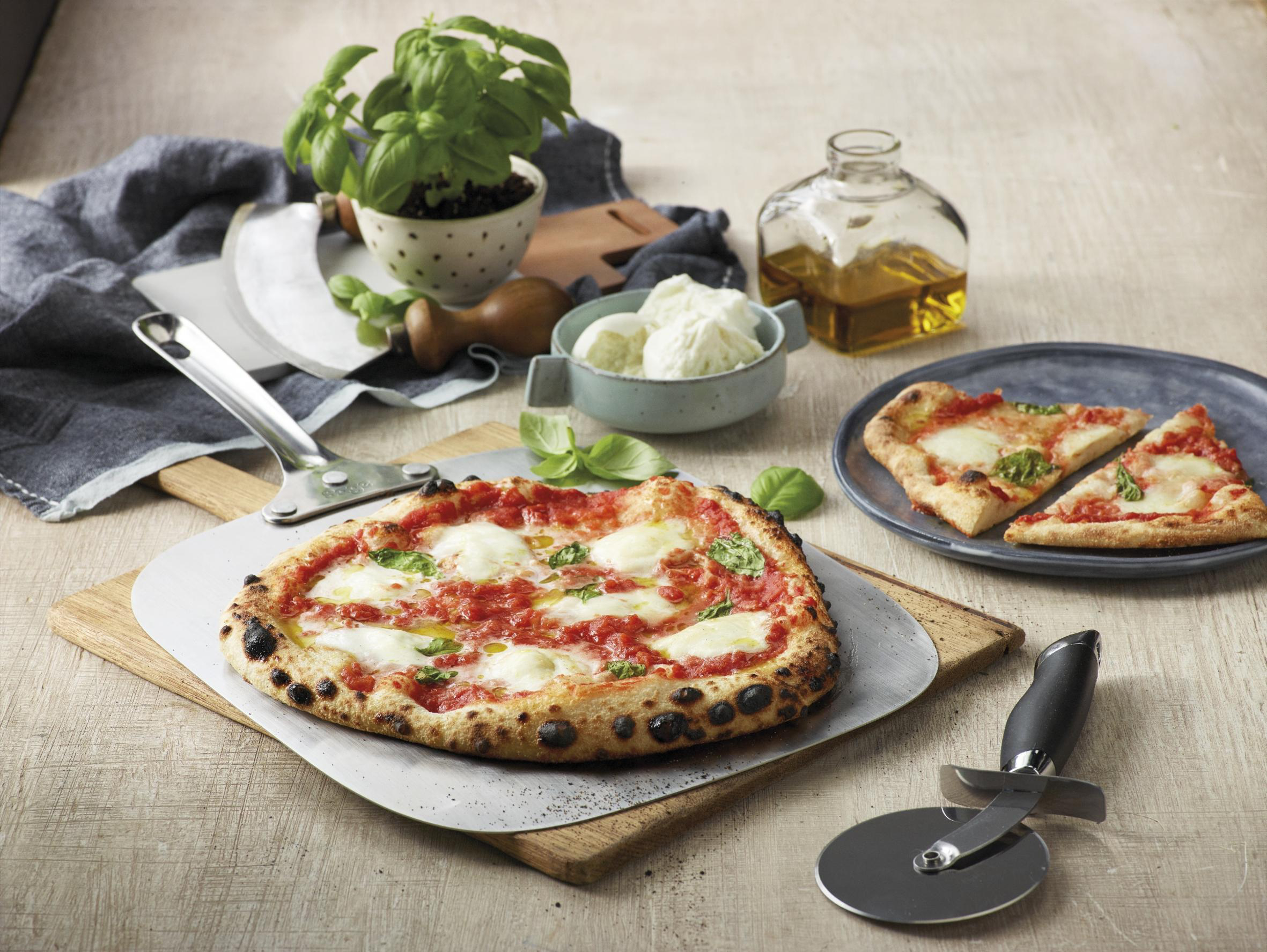 Pizza auf einem Holzbrett, Pizzaschneider, Basilikum, Olivenöl und zwei Pizzastücke.