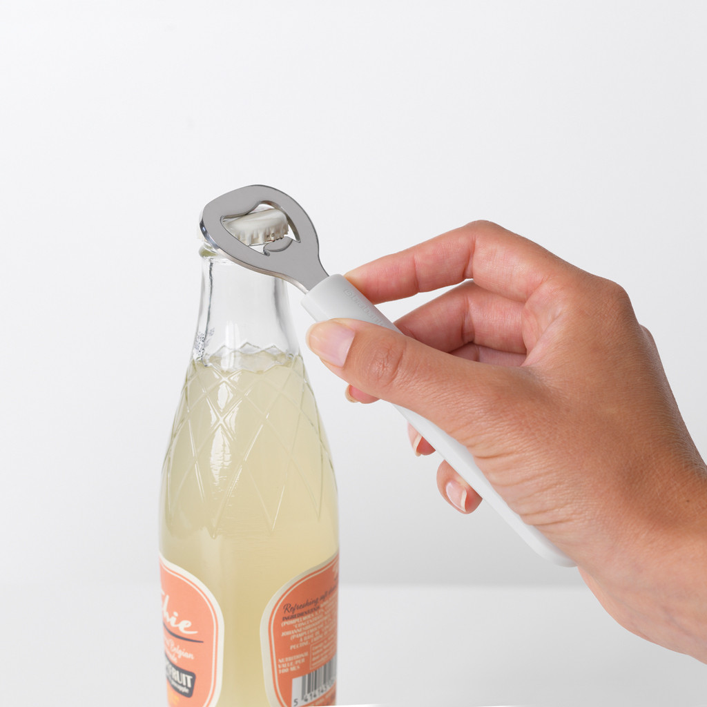 A hand uses a bottle opener to open a bottle of soda. The bottle is orange labeled.
