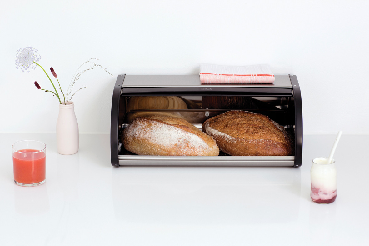 Une boîte à pain sur un comptoir blanc contient du pain. Un verre de jus et du yaourt sont sur les côtés.