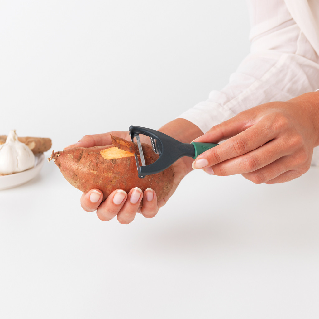 A person peels a sweet potato with a peeler. A garlic clove and a ginger root are in the background on a white plate.