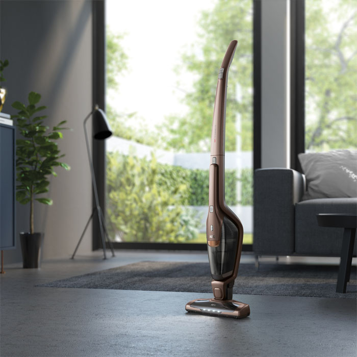 Ein bronzefarbener Akku-Staubsauger steht in einem Wohnzimmer mit Sofa, Lampe und Blick nach draußen.