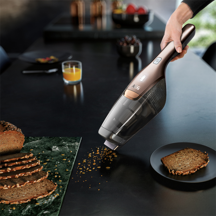 Ein Handstaubsauger reinigt Brotkrümel von einem schwarzen Tisch. Brot, Saft und Obst sind ebenfalls vorhanden.