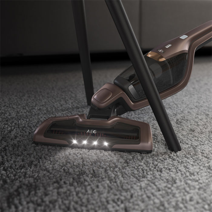 Ein brauner AEG-Staubsauger auf grauem Teppich. Die Lichter am Kopf leuchten. Dunkle Beine und ein dunkler Hintergrund sind sichtbar.
