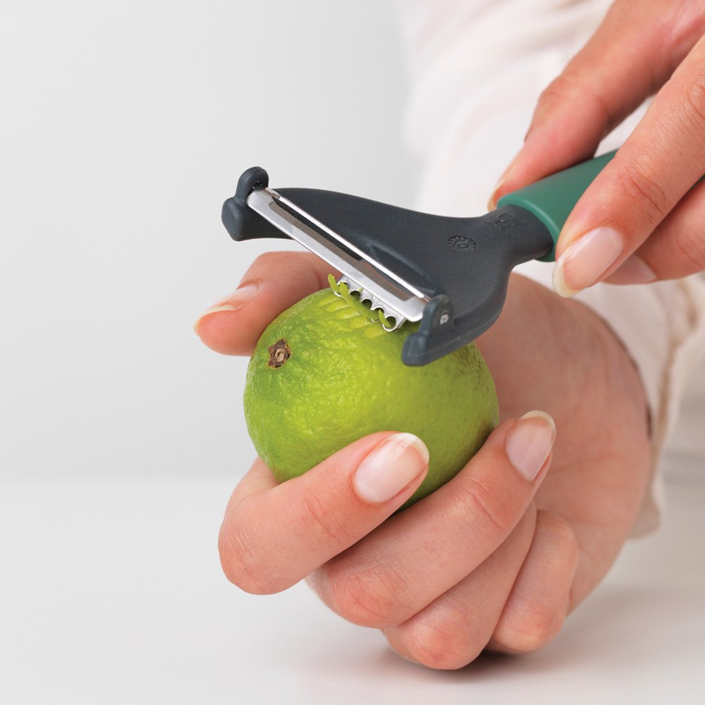 A person peeling a green lime with a peeler. The peeler is dark gray with a green handle. White background.