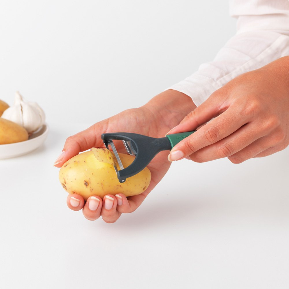 A person peeling a potato with a green and black peeler. Other potatoes and garlic are in the background, on a white surface.