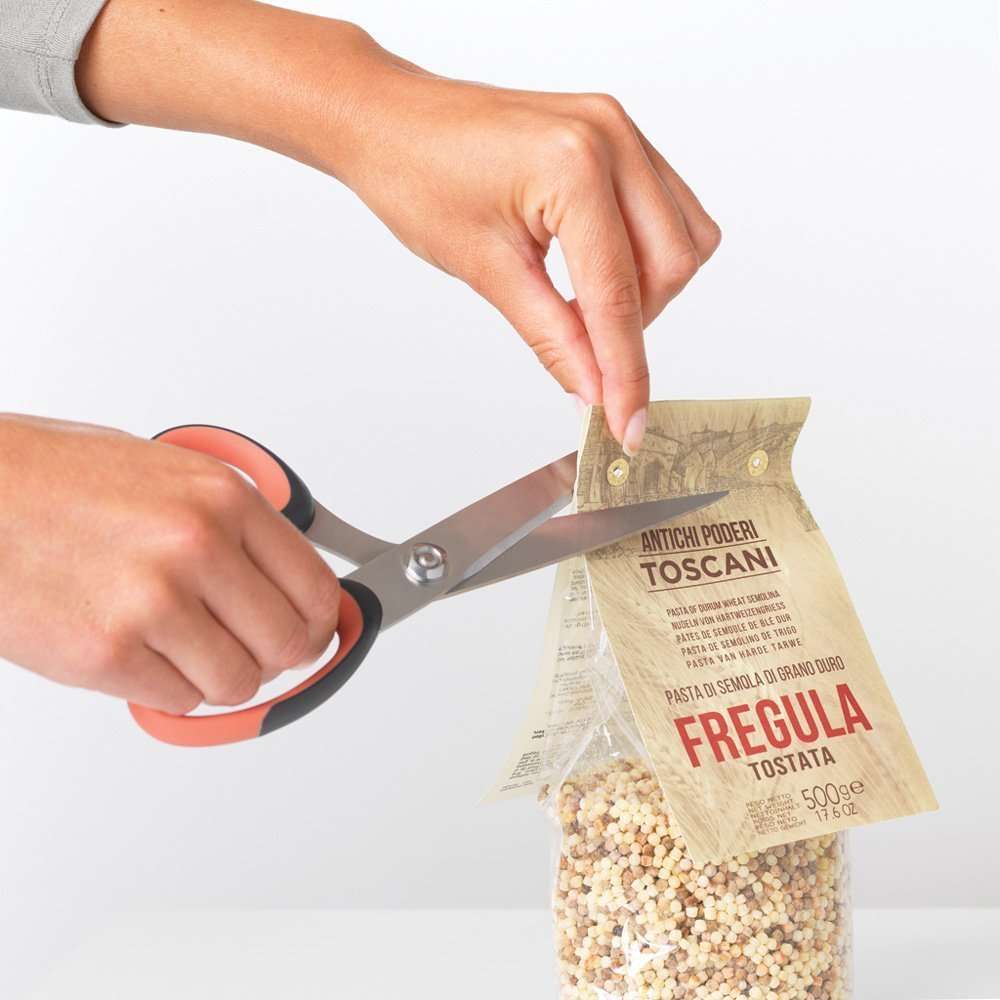 Hands using scissors to cut open a pasta bag labeled "Fregula Tostata". The bag is filled with small, round pasta.