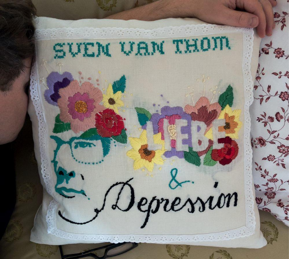Besticktes Kissen mit Blumen und Text: Sven Van Thom, Liebe & Depression. Der Kopf einer Person befindet sich in der linken Ecke.