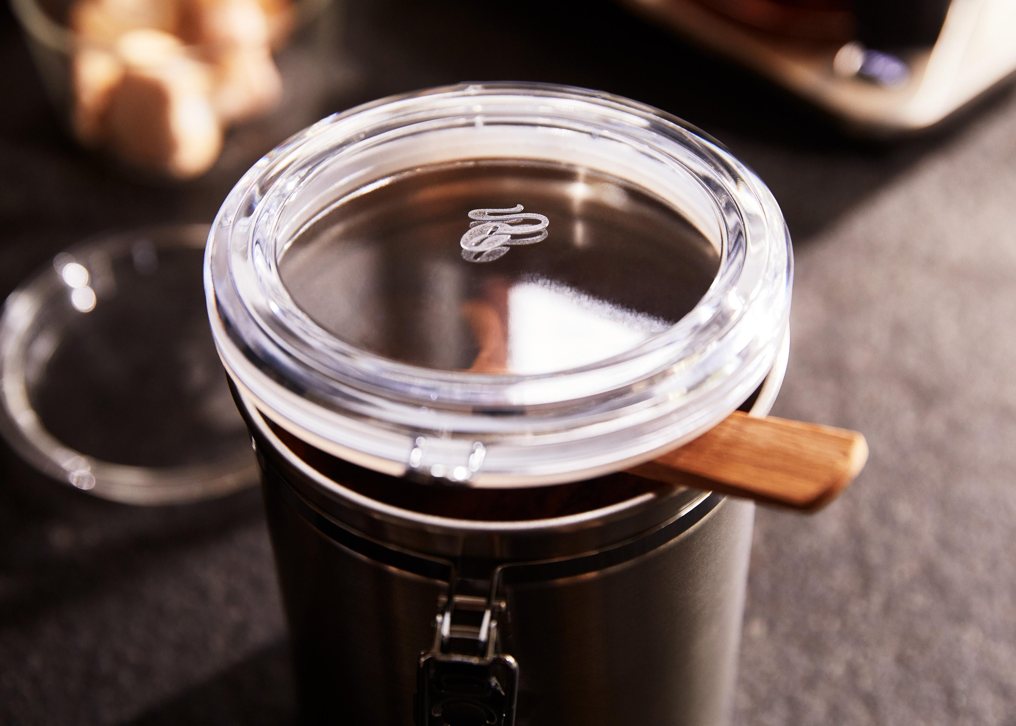 Stahlbehälter mit Deckel, Holzlöffel innen. Kaffeebohnen, dunkle Oberfläche.