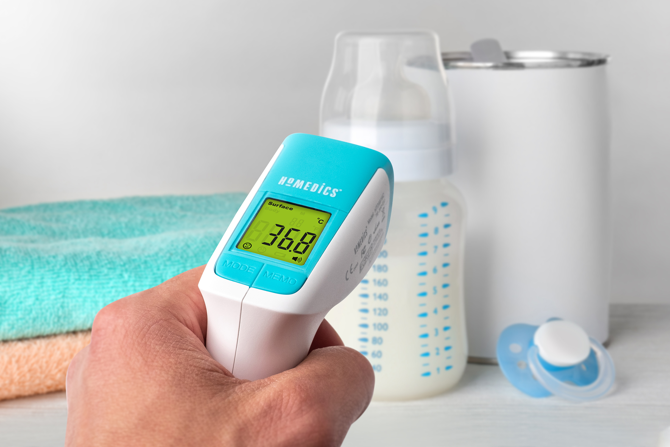 A hand holding a blue and white digital thermometer near baby items.